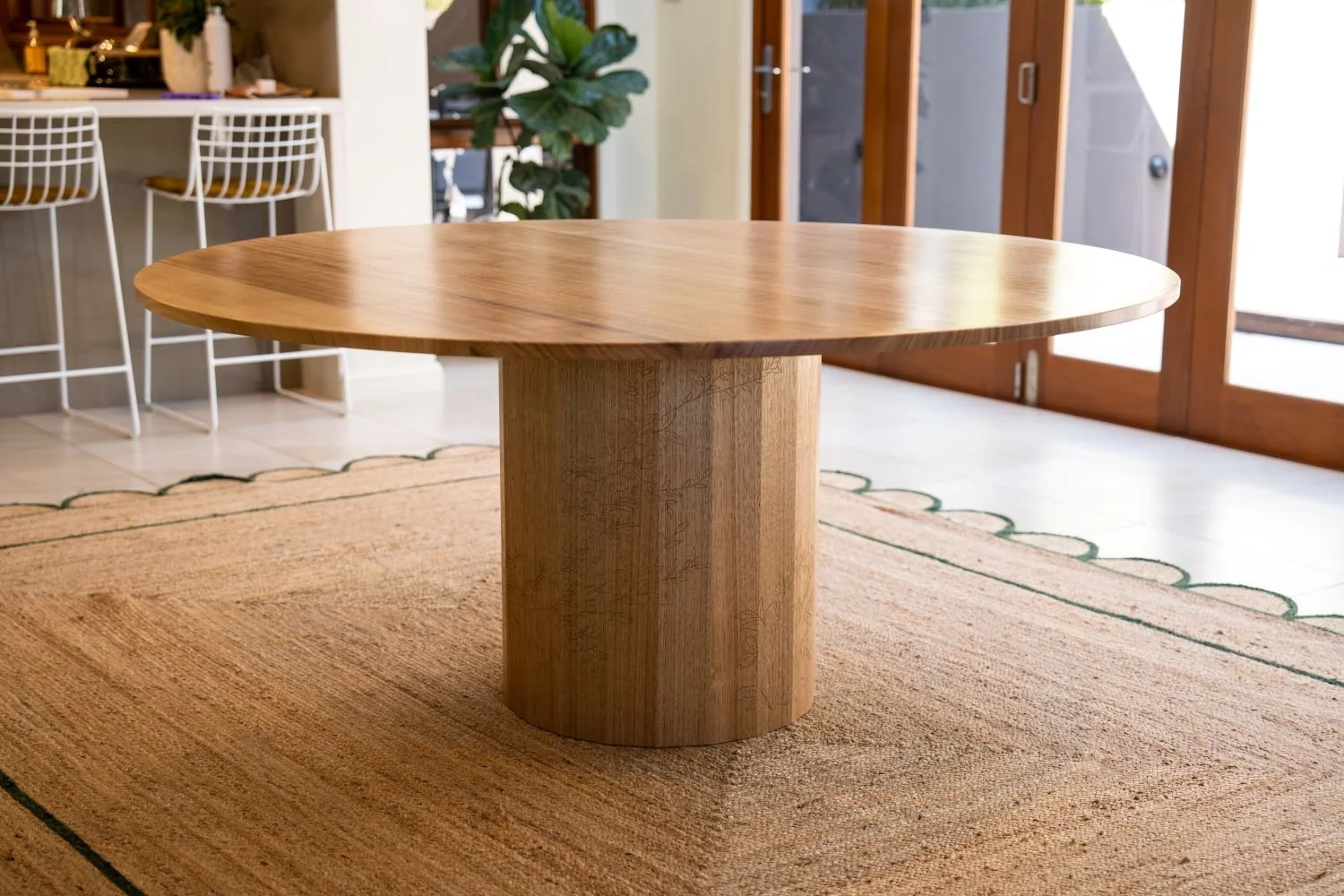 A round wooden table with a thick pedestal base on a woven rug in a bright room with large glass doors and a plant in the background.