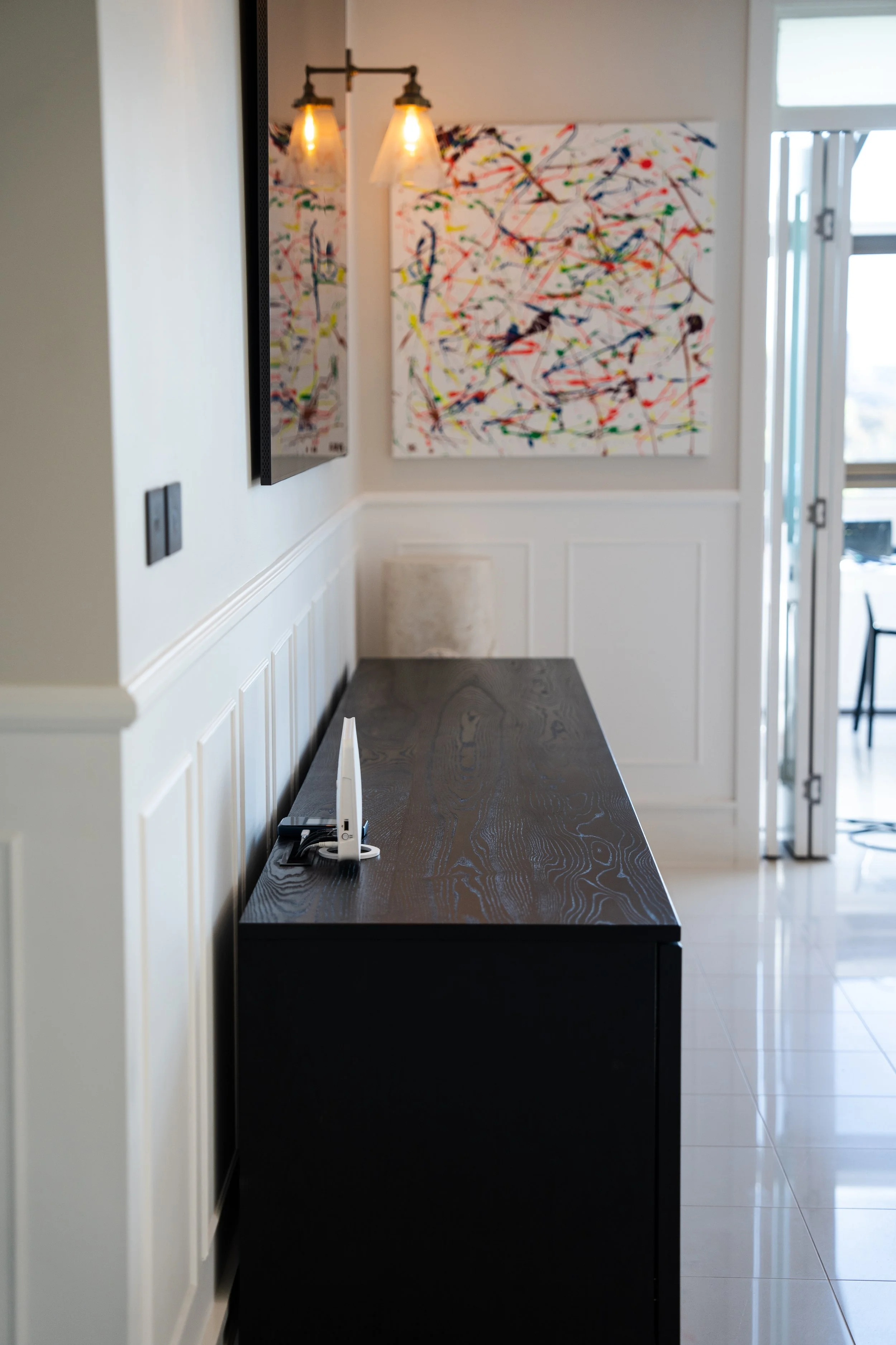 A modern interior with a black wooden sideboard against a white paneled wall, decorated with colorful abstract paintings, a wall-mounted lamp with Edison bulb fixtures, and a partially visible Wi-Fi router on the sideboard.