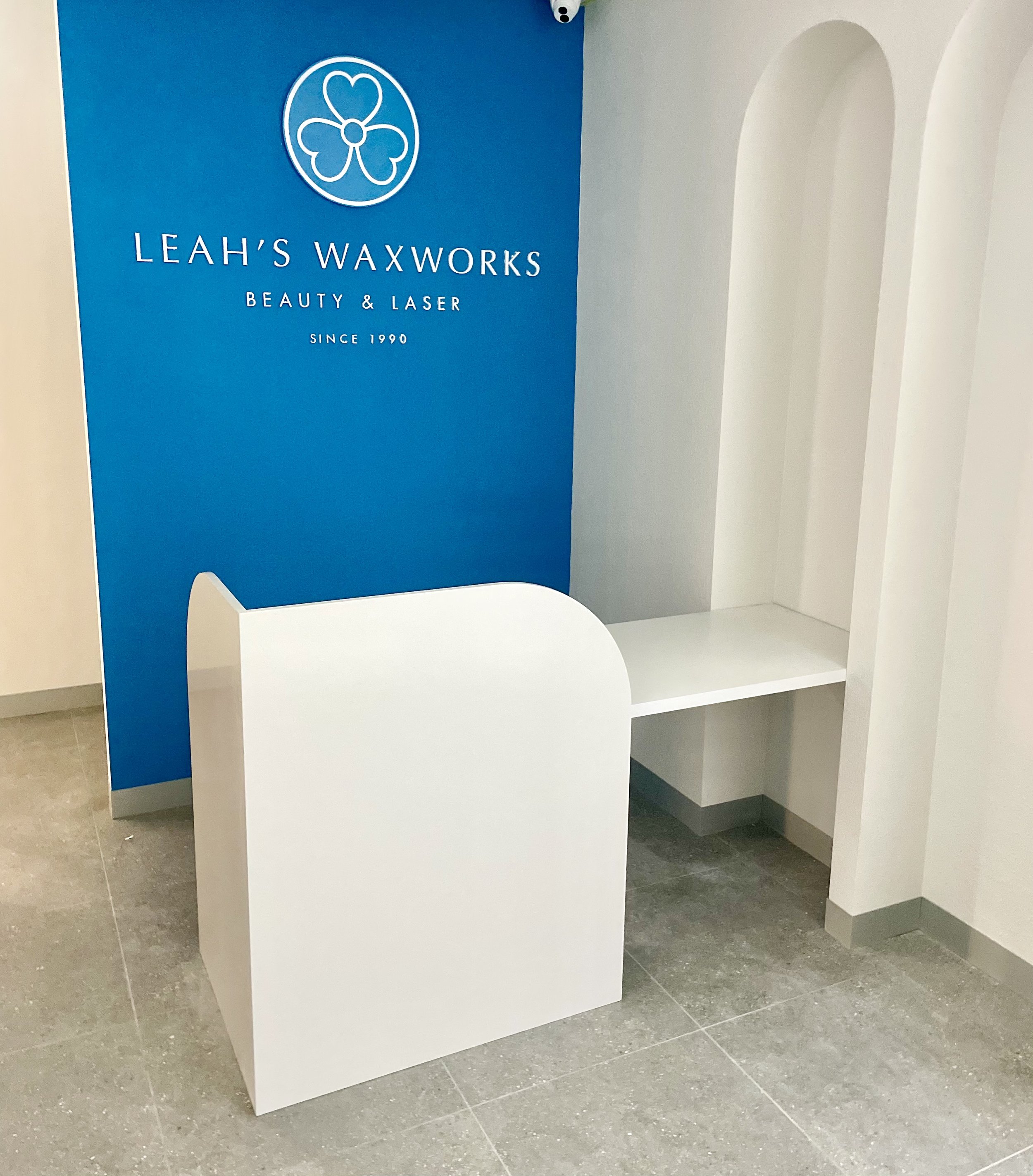 Interior of a beauty and laser salon with branding on a blue wall, displaying a white reception desk, a wall with three white arches, and the salon's logo featuring a flower and the name Leah's Waxworks.