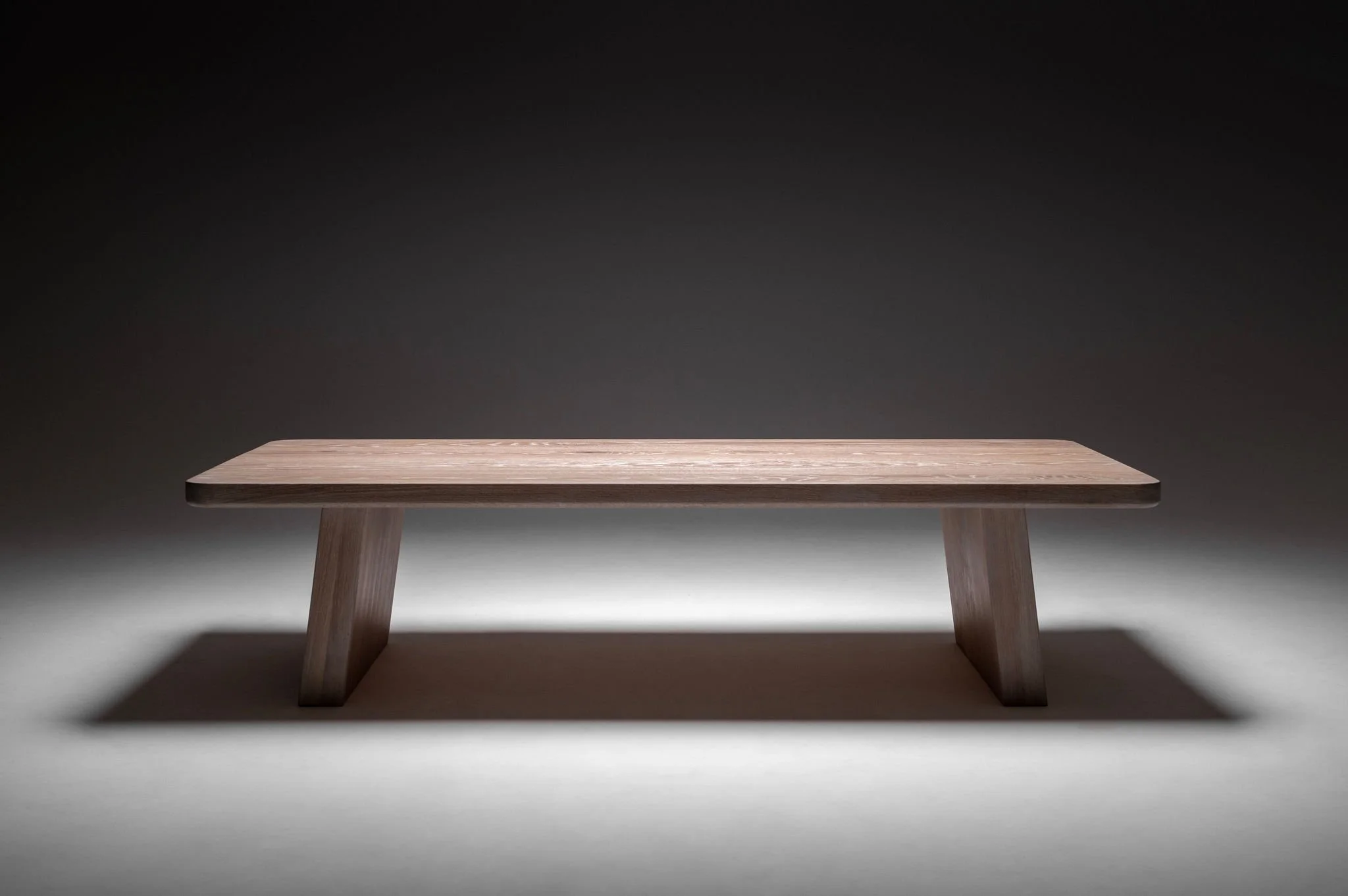 A simple wooden bench with a flat rectangular top and two angled legs, set against a dark background with spot lighting.