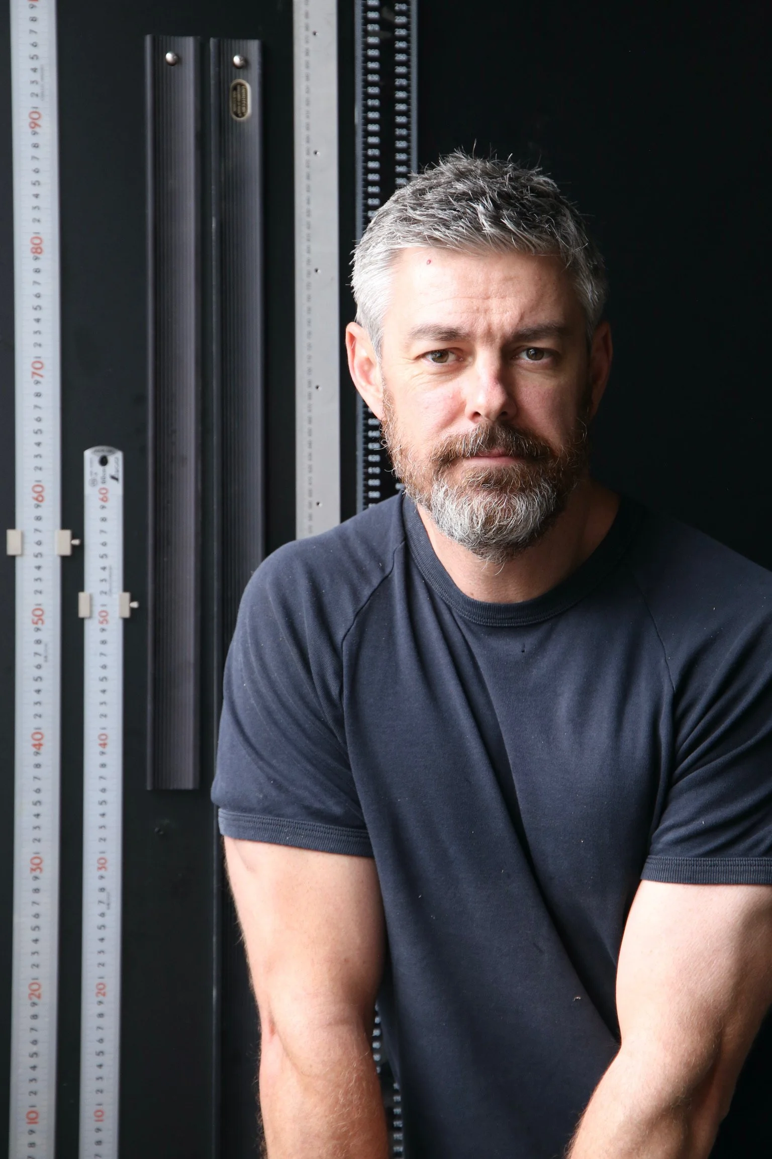 A man with gray hair and a beard, wearing a black T-shirt, sitting next to a background of measurement tools, including rulers and a grid or slider, against a black backdrop.