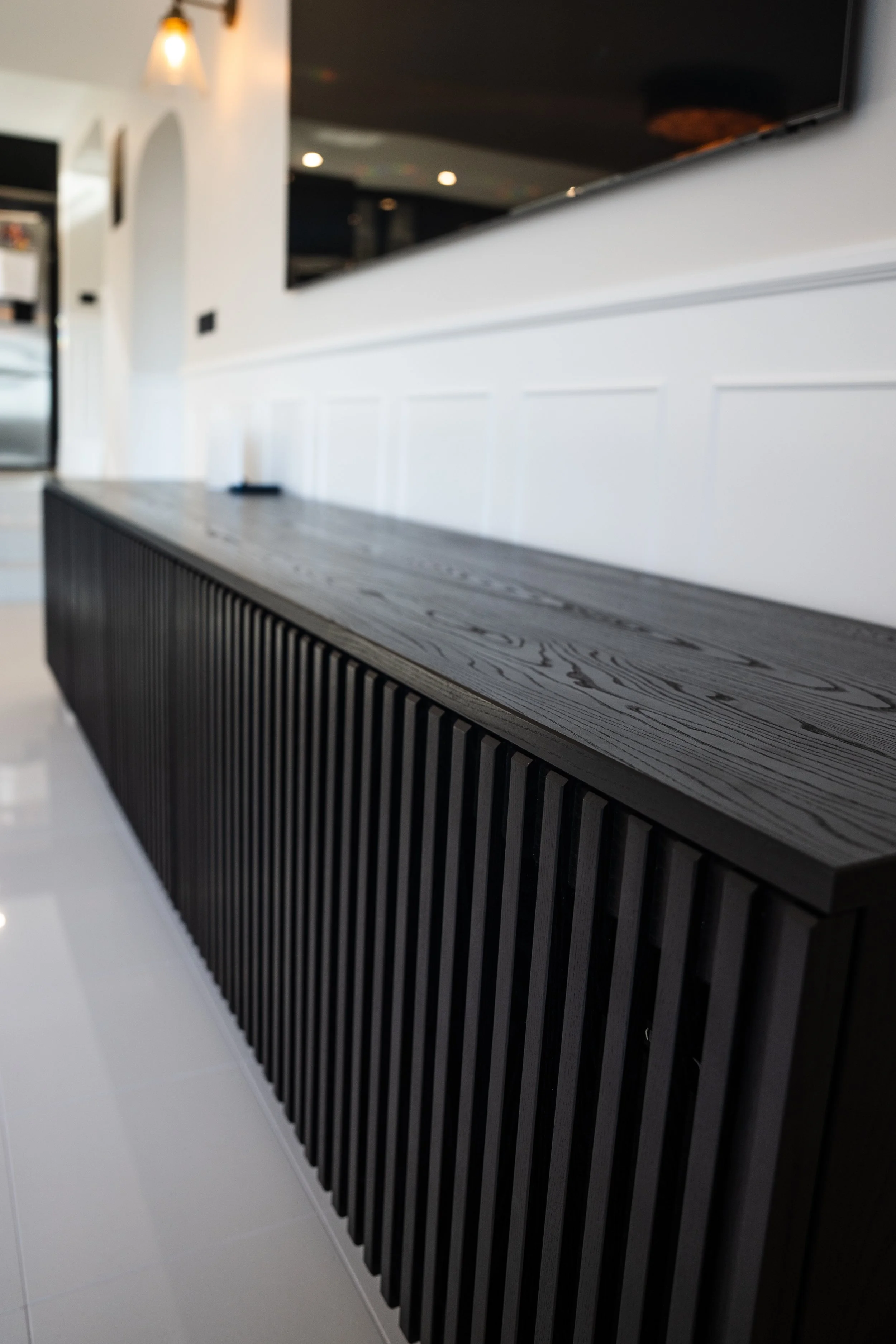 Close-up of a modern black wooden bench with vertical slats in a well-lit interior space, with a white wall and reflections on glossy flooring in the background.