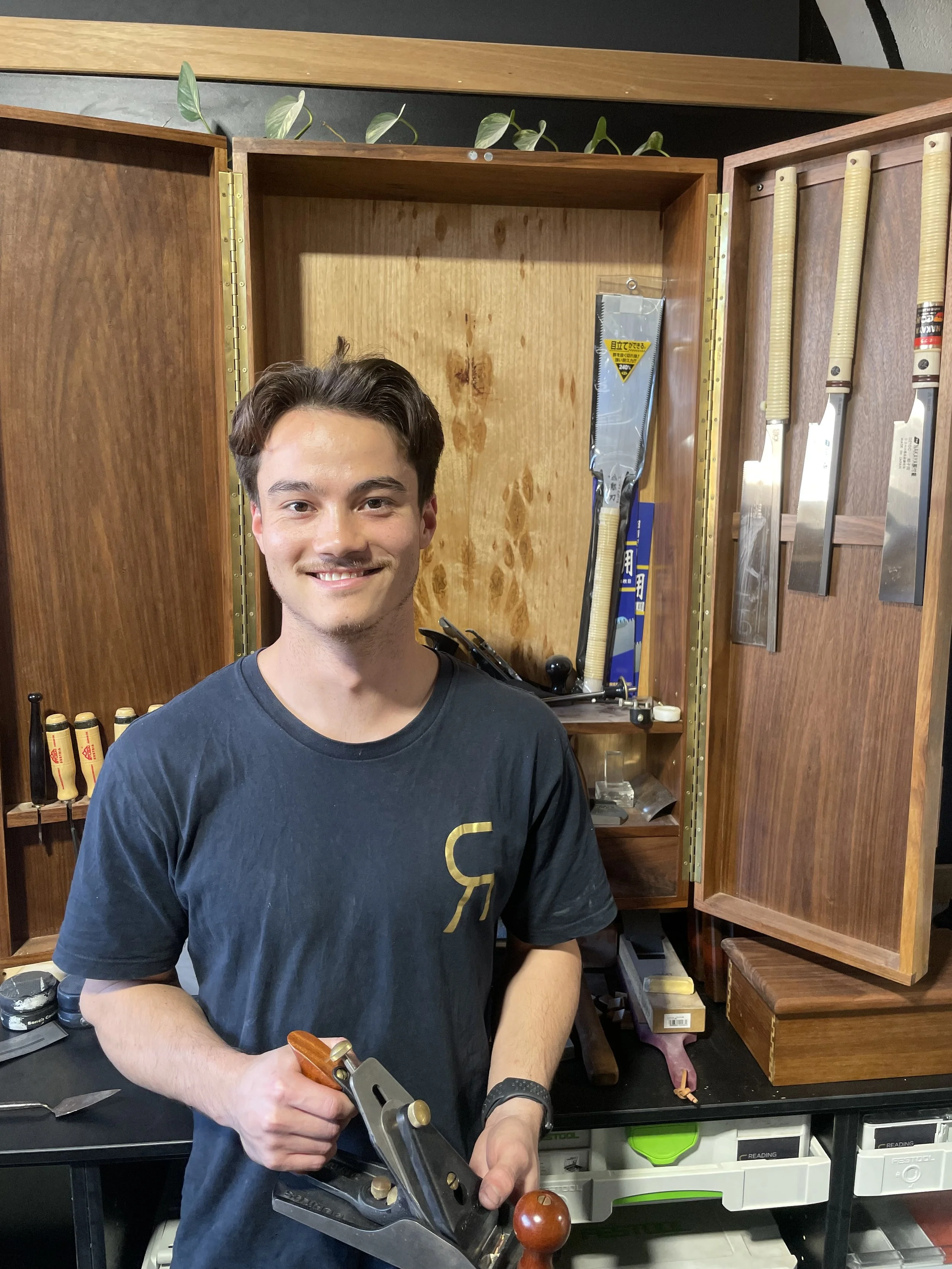Young man smiling while holding a hand plane in a woodworking workshop, with tools and woodworking supplies in the background.