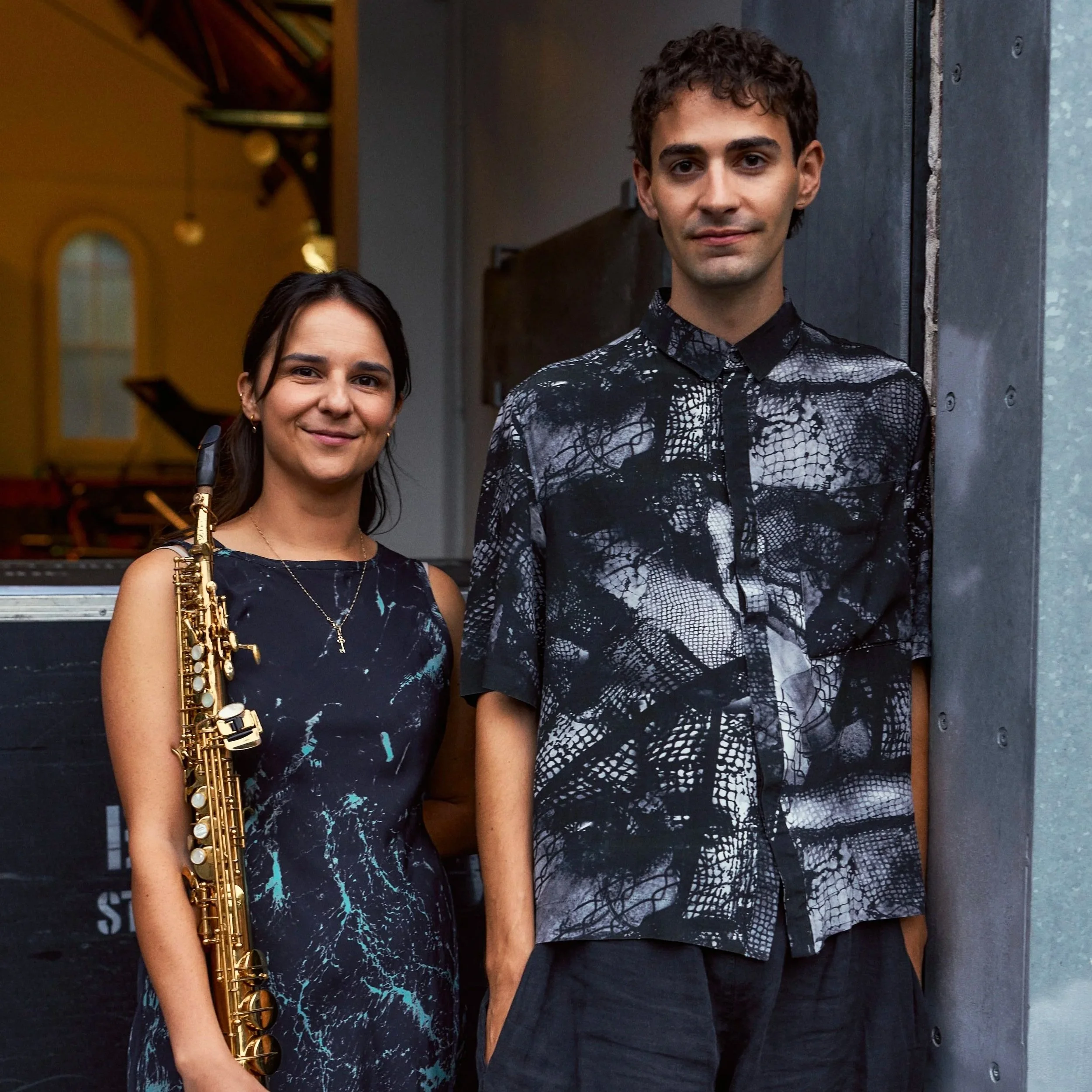 A woman holding a saxophone smiling and a man standing next to her, both dressed in black with snake skin pattern shirts, standing outside near a building.