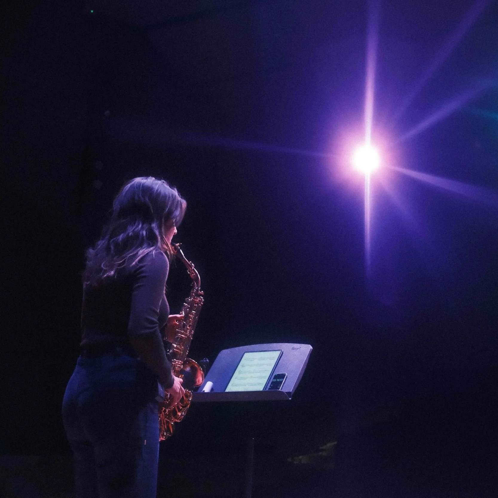 A woman with long, wavy hair playing a saxophone on a dark stage, illuminated by a bright purple light, with a music stand displaying sheet music and a digital device.