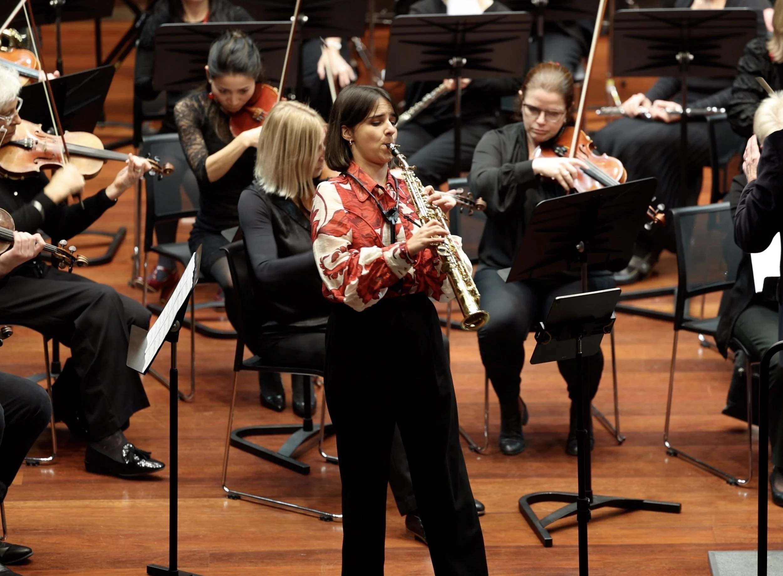 A woman playing the saxophone during an orchestra performance, with other musicians playing violins and woodwinds in the background on a wooden stage.