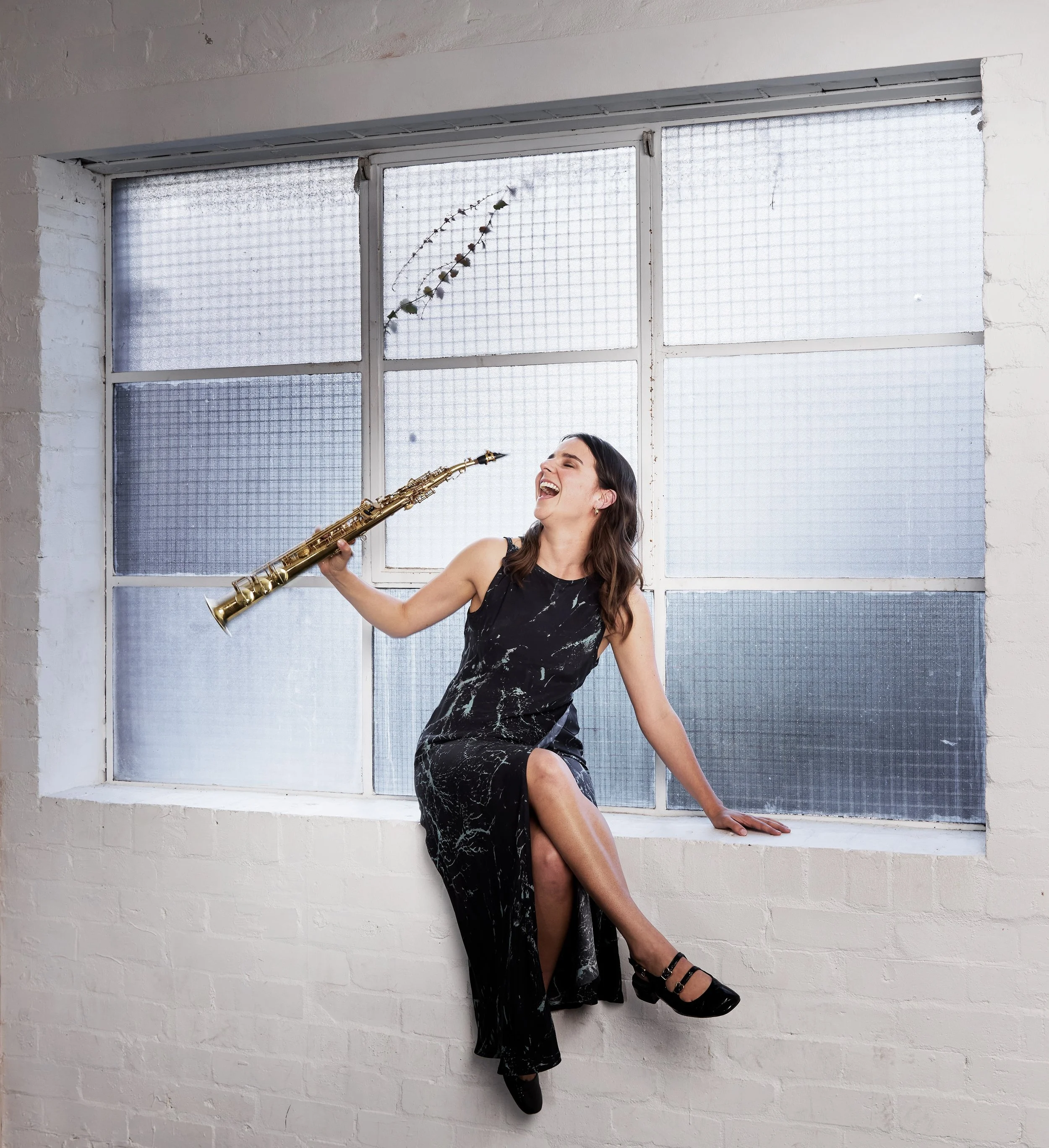 A woman with long dark hair, wearing a black marble-patterned dress, is sitting on a window sill, laughing and playing a golden saxophone. The window has frosted glass panes and is set in a white brick wall.