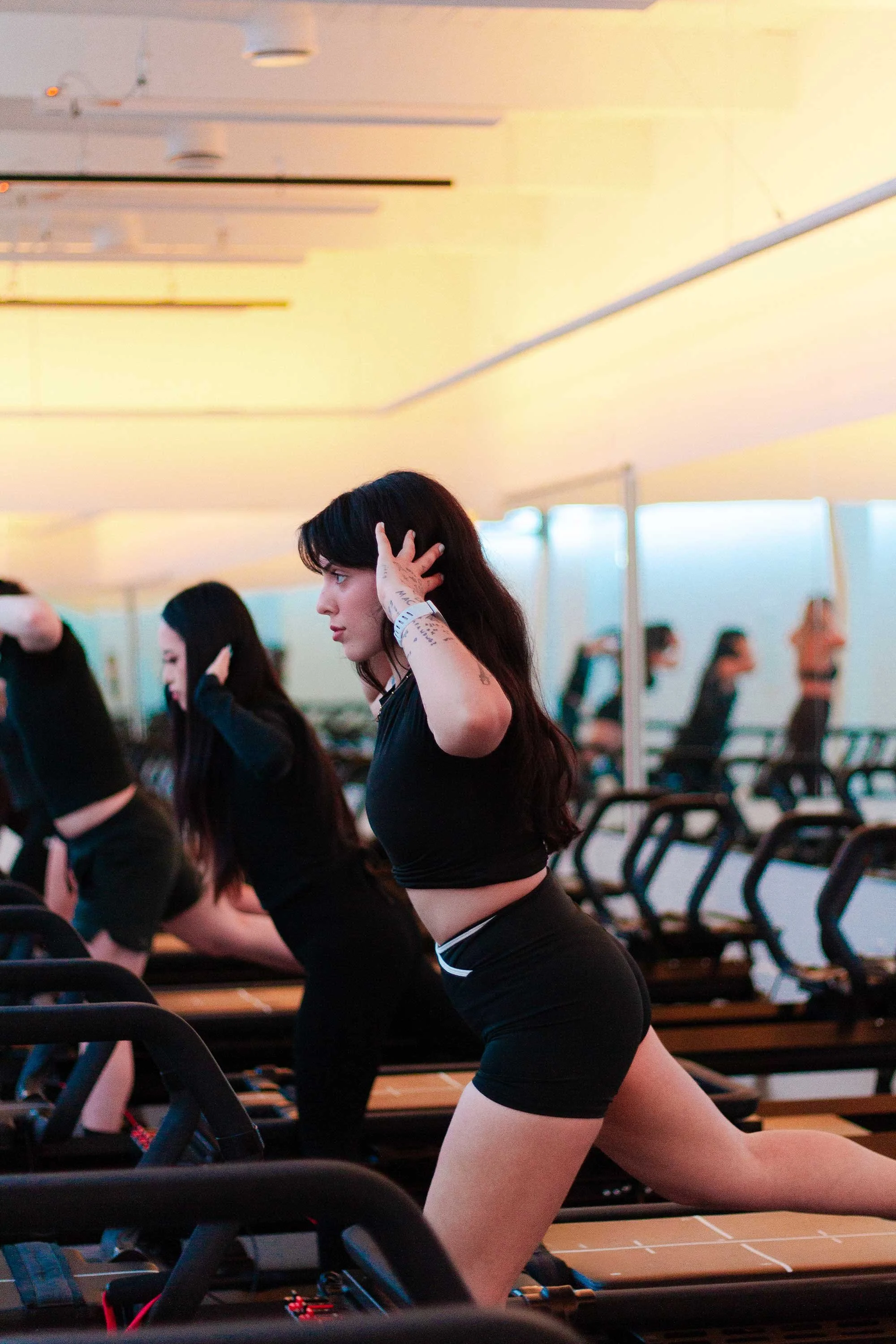 A young woman with black hair doing a lunging exercise on a rowing machine in a gym, surrounded by other people also exercising.