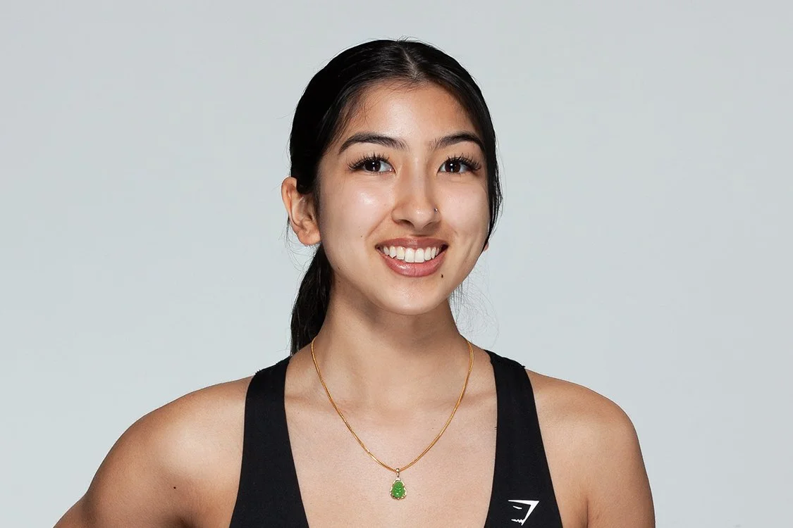 Young woman smiling, black athletic tank top, gold necklace with green pendant, black hair in a ponytail, light background.