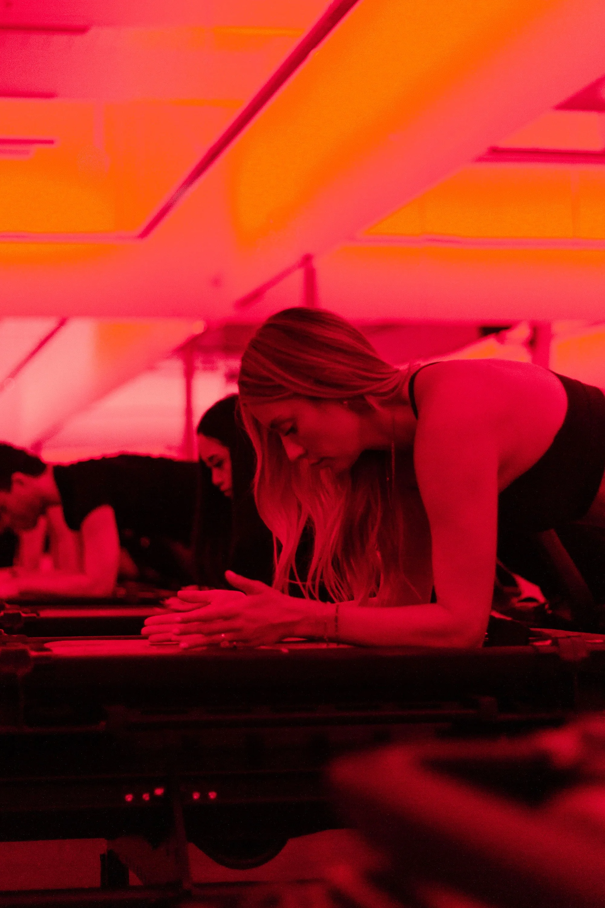 A woman with long hair is doing a plank exercise on a yoga mat in a dimly lit room with red hue lighting.