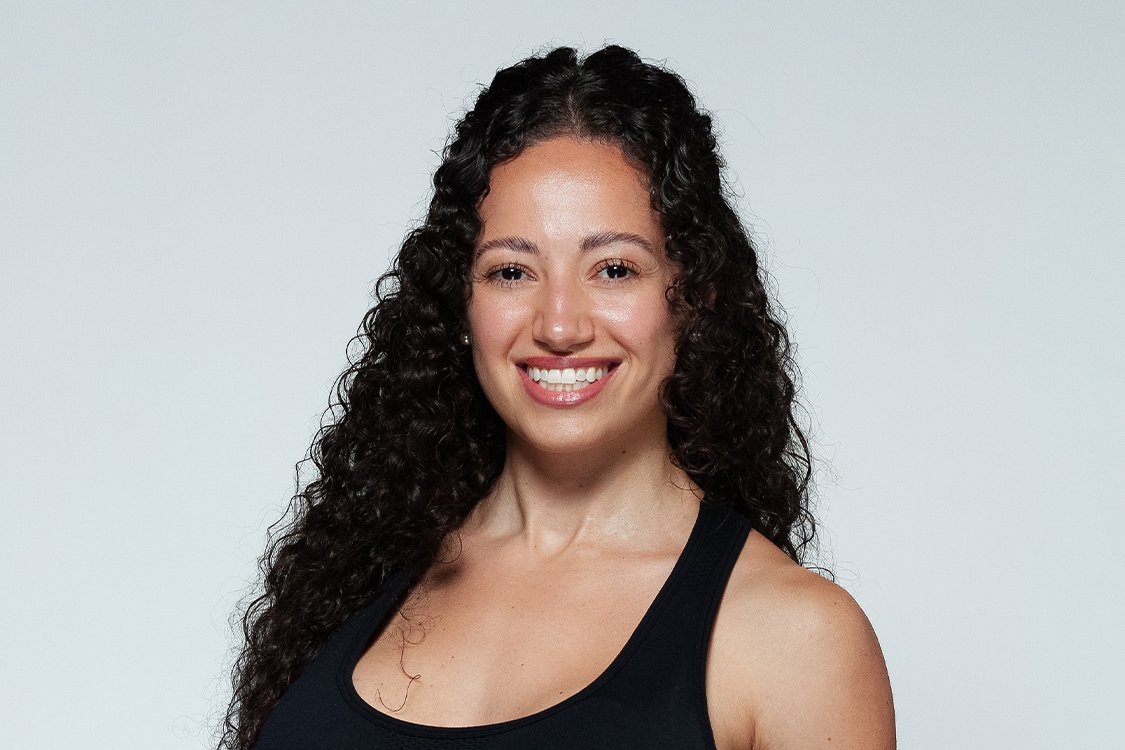 A woman with long, curly dark hair smiling at the camera, wearing a black tank top against a plain white background.