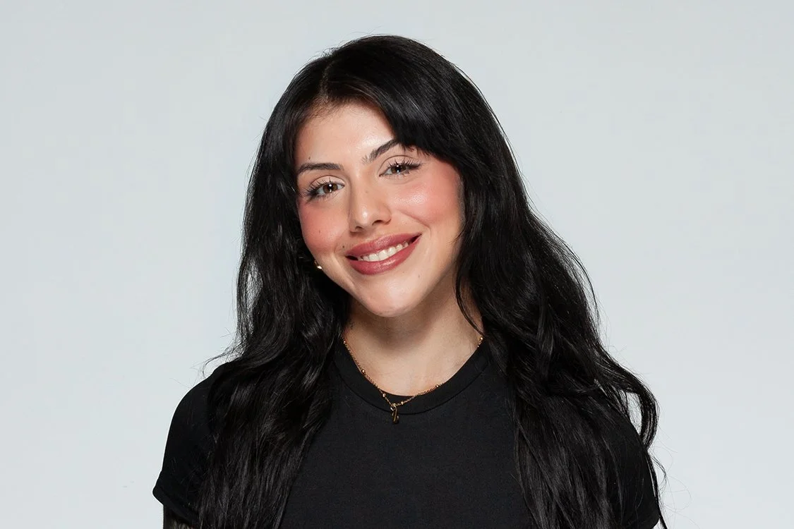 Young woman with dark wavy hair, smiling, wearing a black shirt and a delicate gold necklace, standing against a light gray background.