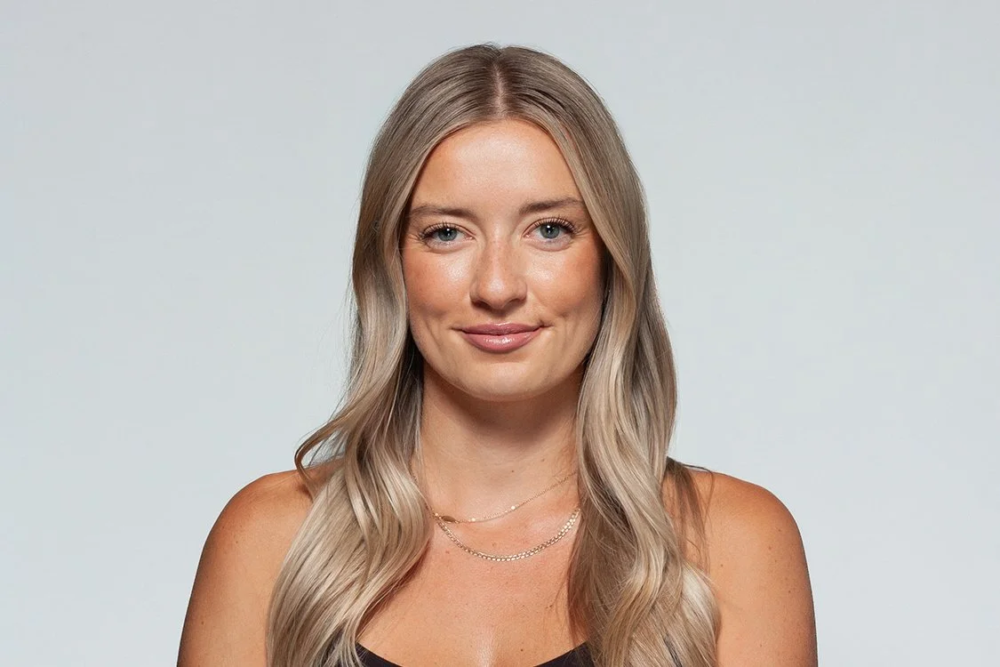 A young woman with long, wavy blonde hair, wearing a black sleeveless top and layered necklaces, smiling softly at the camera against a plain light-colored background.