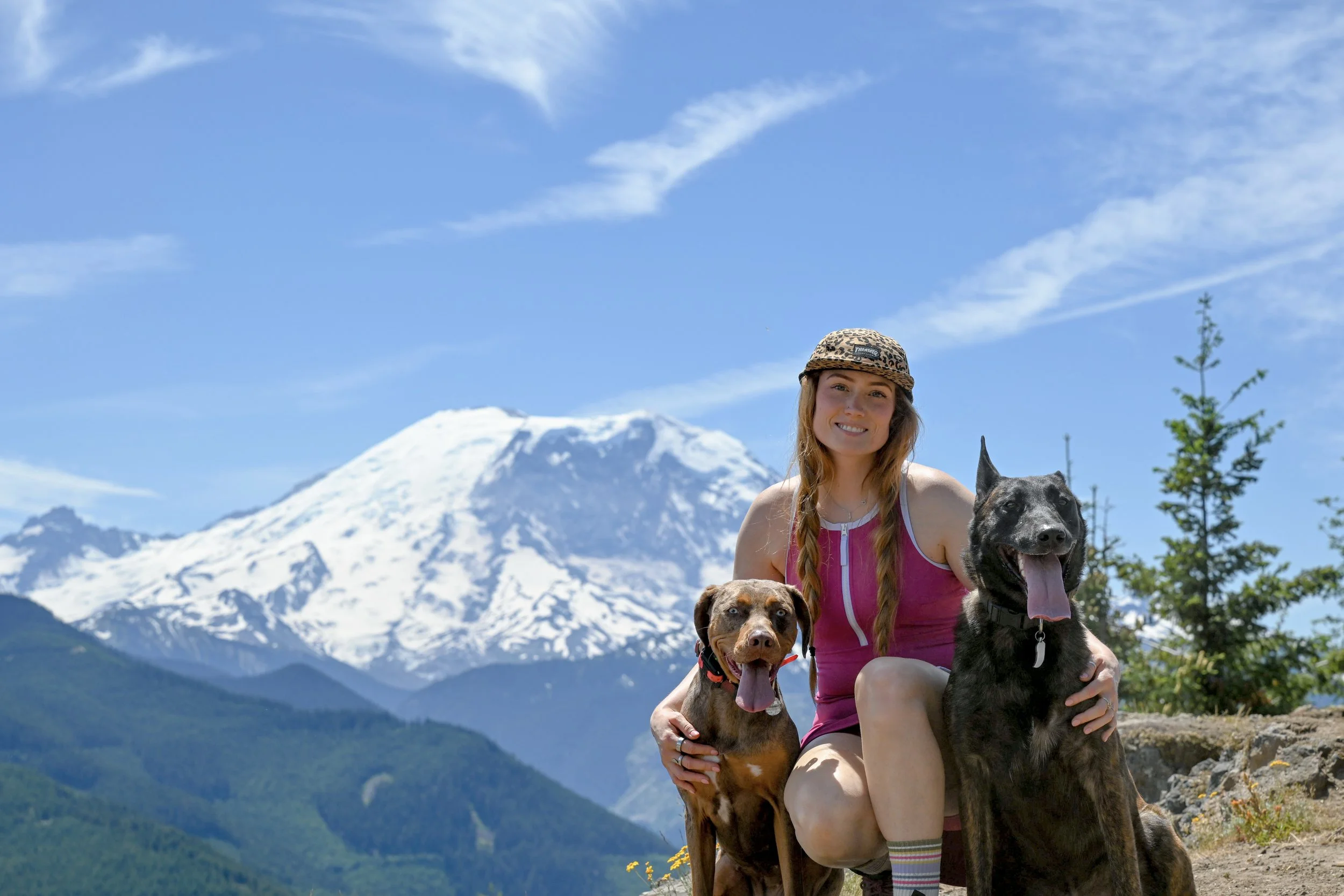 A woman with long hair, wearing a pink tank top and a leopard print hat, kneeling outdoors with two dogs in front of a snow-capped mountain and a bright blue sky.