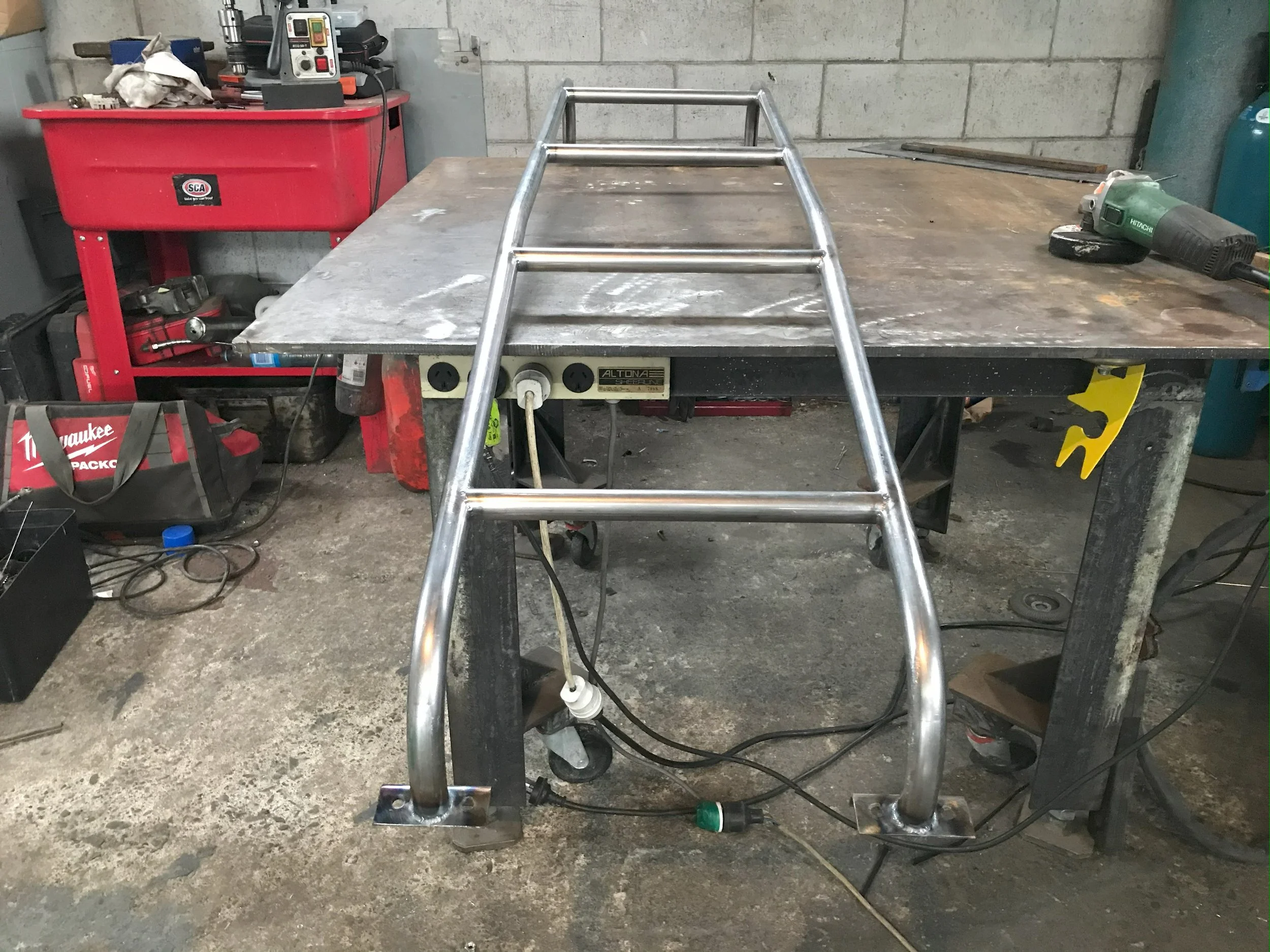 A metal frame being welded on a workbench in a workshop. Tools and equipment are visible in the background, including a red tool cabinet and a handheld power tool on the right.