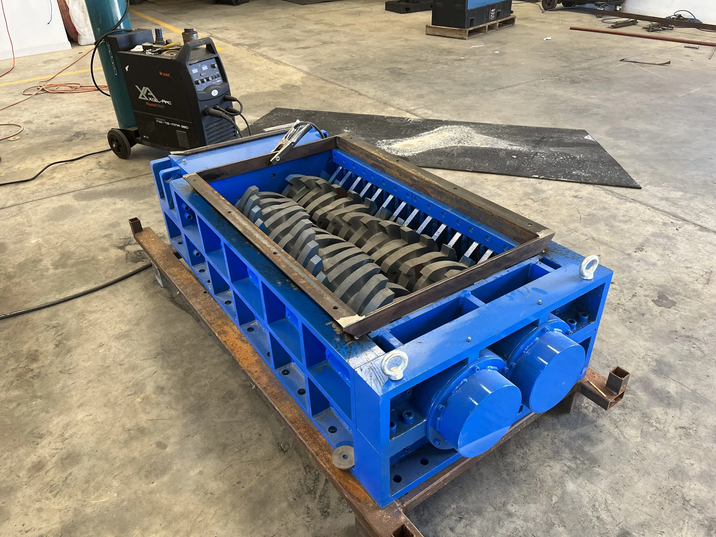 A blue industrial machine with a large rotary roller in the foreground, connected to a black welding machine in a warehouse or workshop setting. There are tools and equipment scattered around on the ground and on nearby pallets.