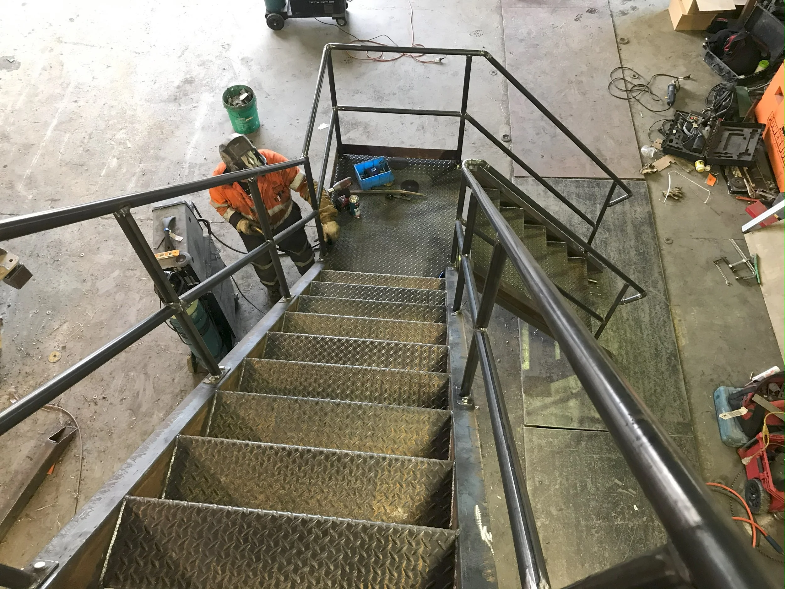 Looking down from the top of a metal staircase in a workshop or garage, with tools and equipment scattered on the concrete floor.