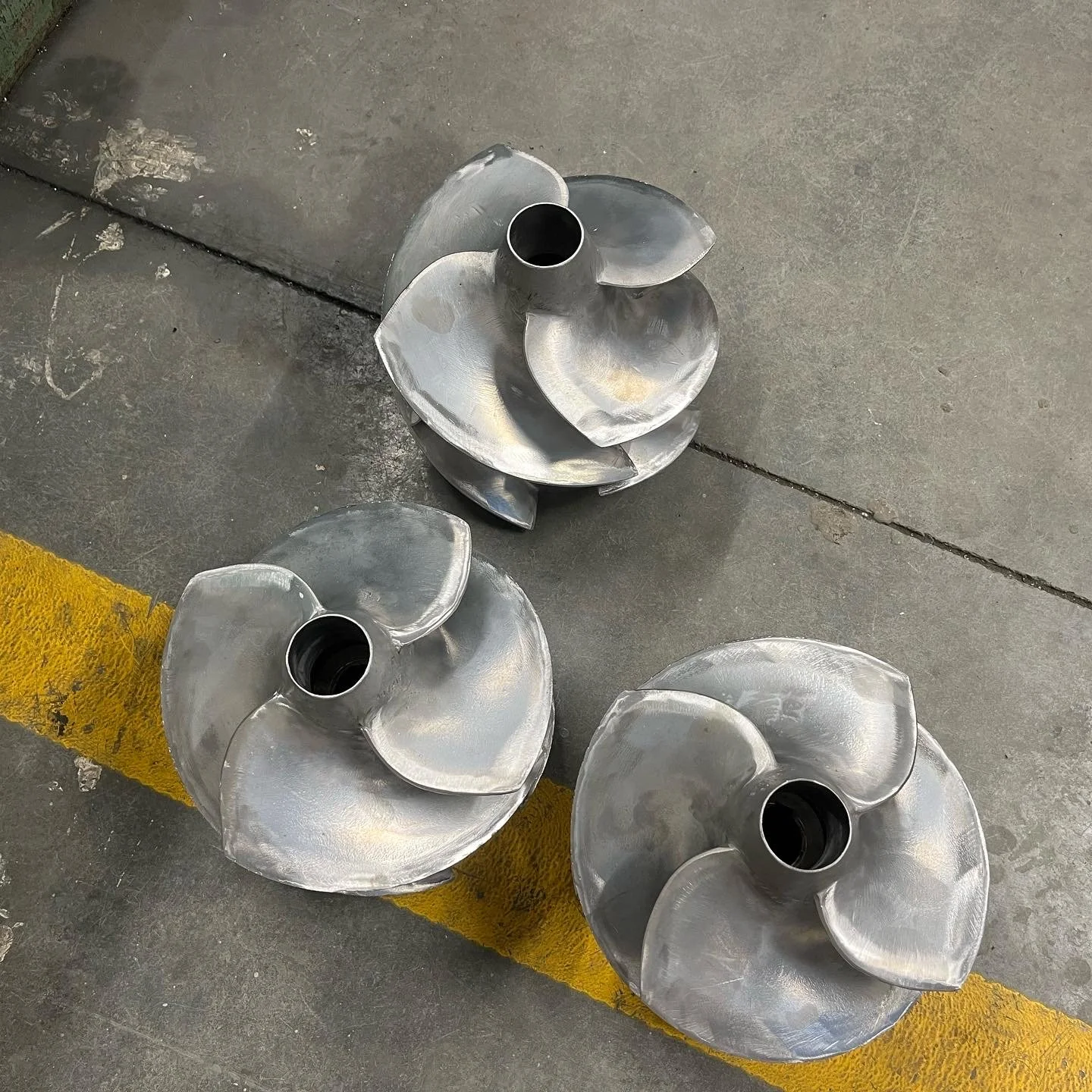 Three metallic boat propellers with sharp blades, lying on a concrete floor near a yellow safety line.