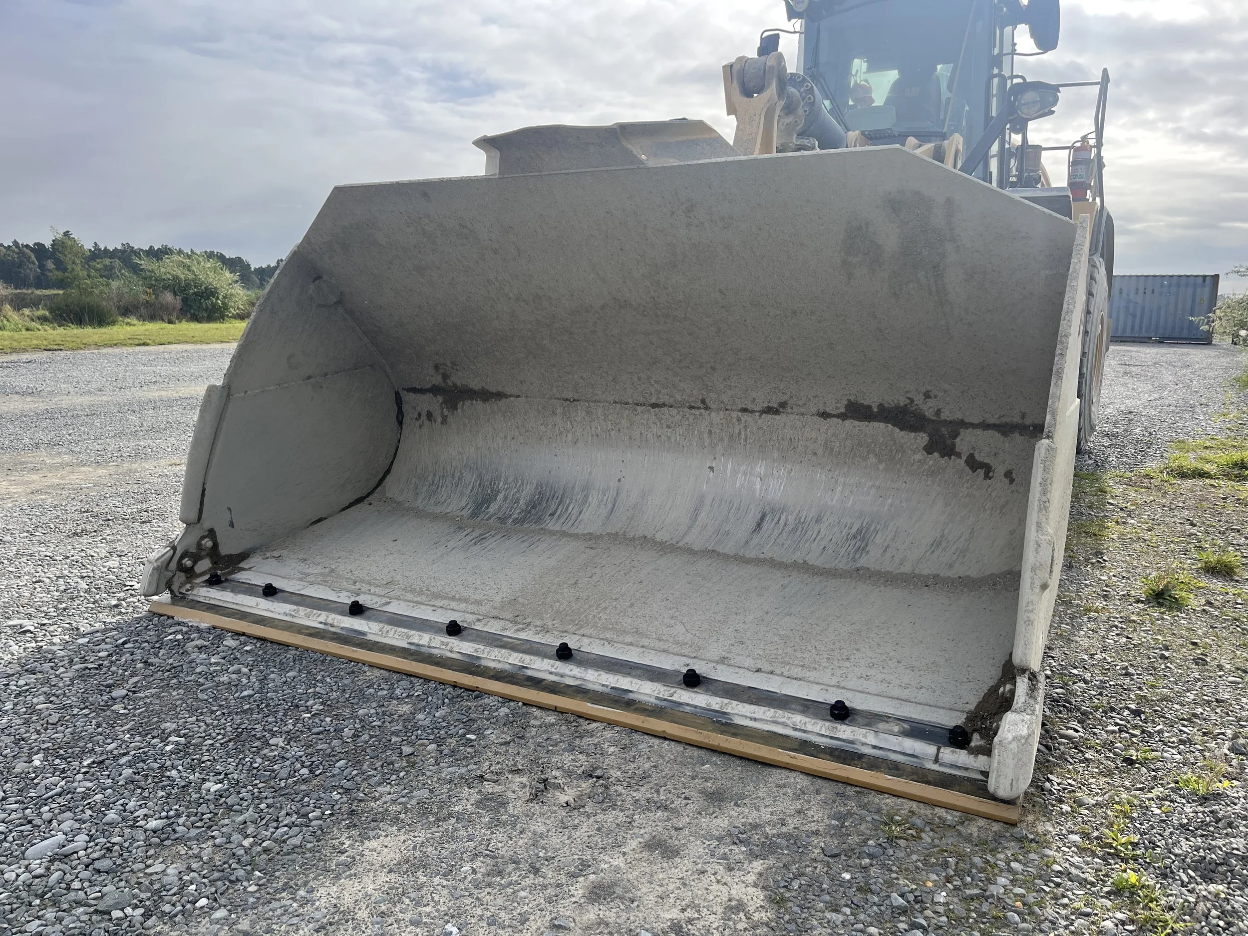 A large construction vehicle with a front blade for grading or pushing earth, parked on a gravel surface outdoors with a cloudy sky overhead.
