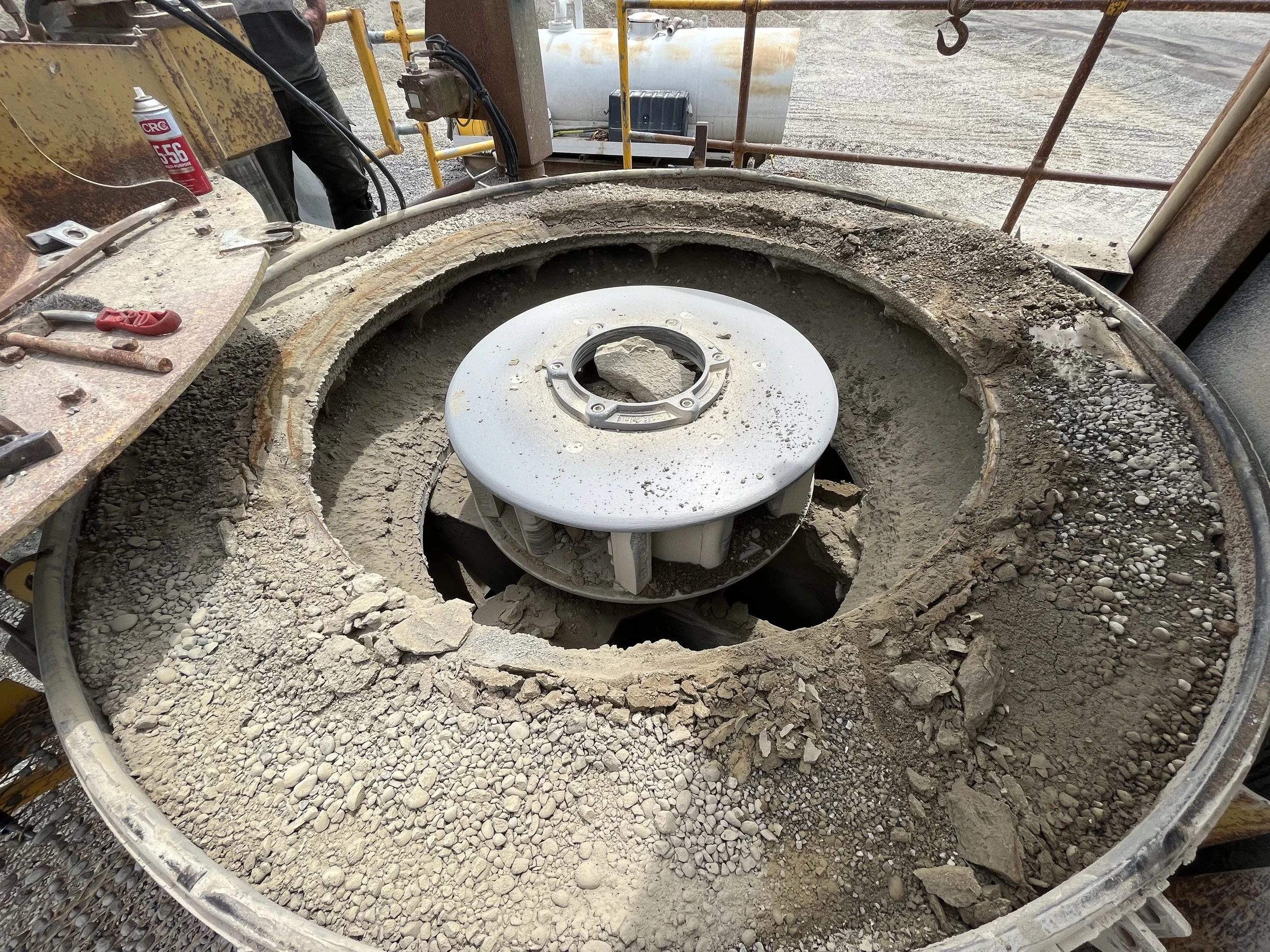 Construction site with a large circular foundation opening filled with gravel and dirt, surrounded by scaffolding and construction tools, with an industrial liquid storage tank in the background.
