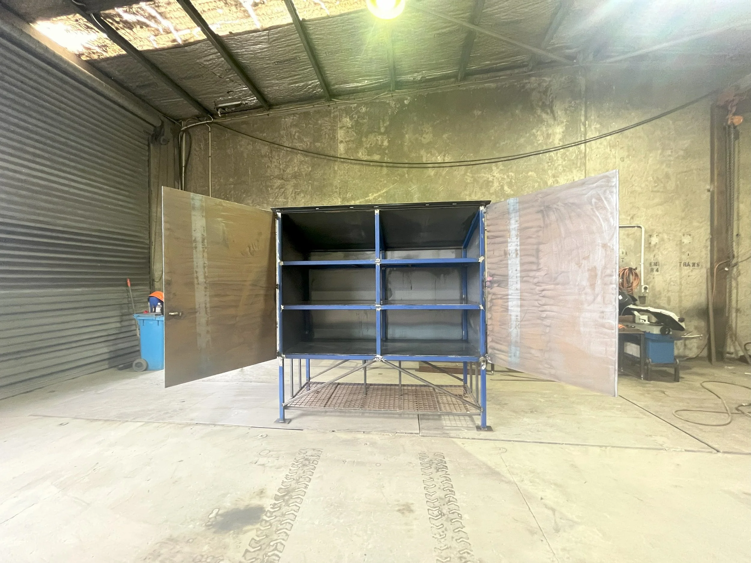 Interior view of a warehouse with a blue shelving unit in the center, a trash can, and tire tracks on the concrete floor. The walls are made of corrugated metal and concrete, with various equipment and boxes visible.
