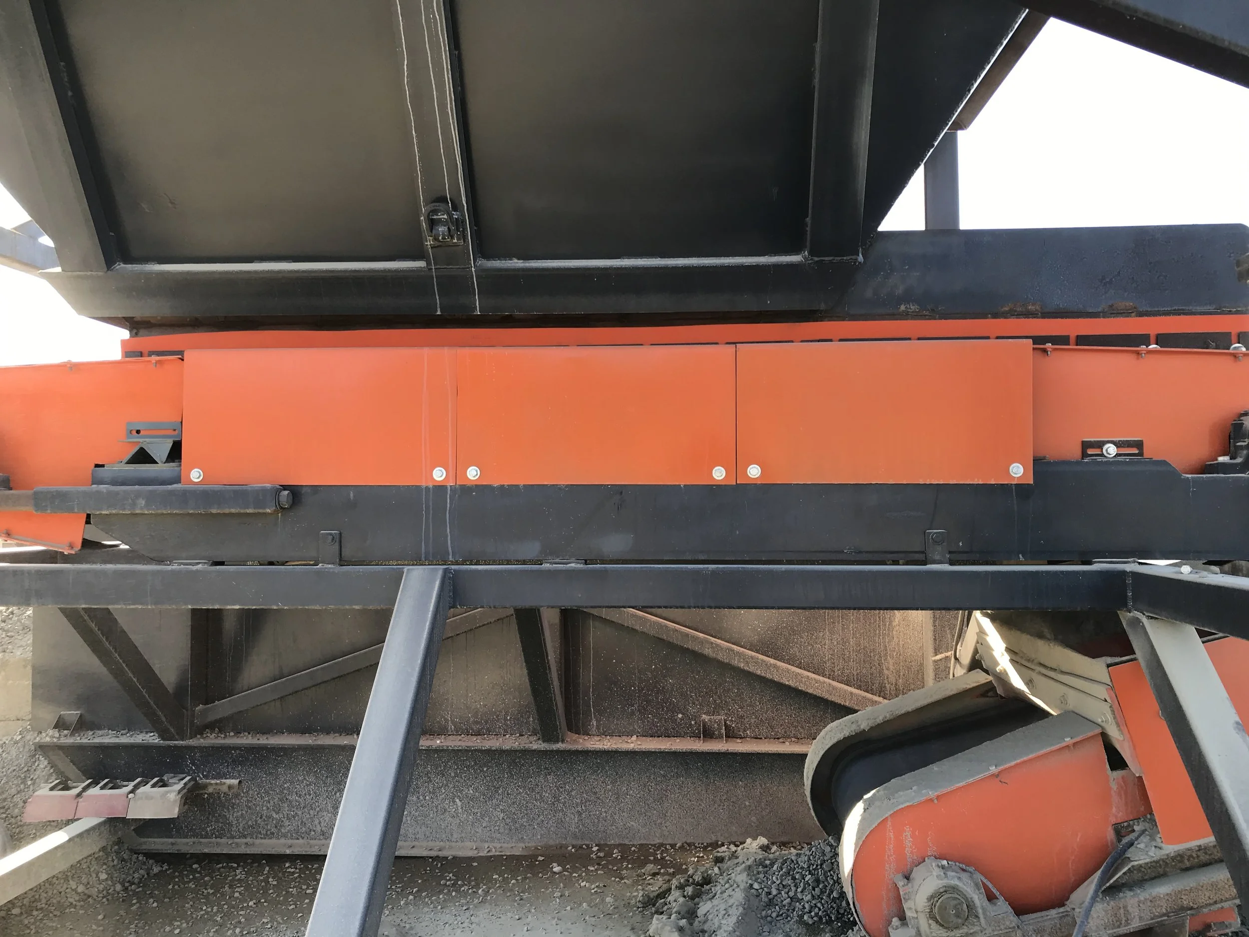 Close-up view of construction equipment with black and orange metal parts, beams, and bolts, on a gravel surface.