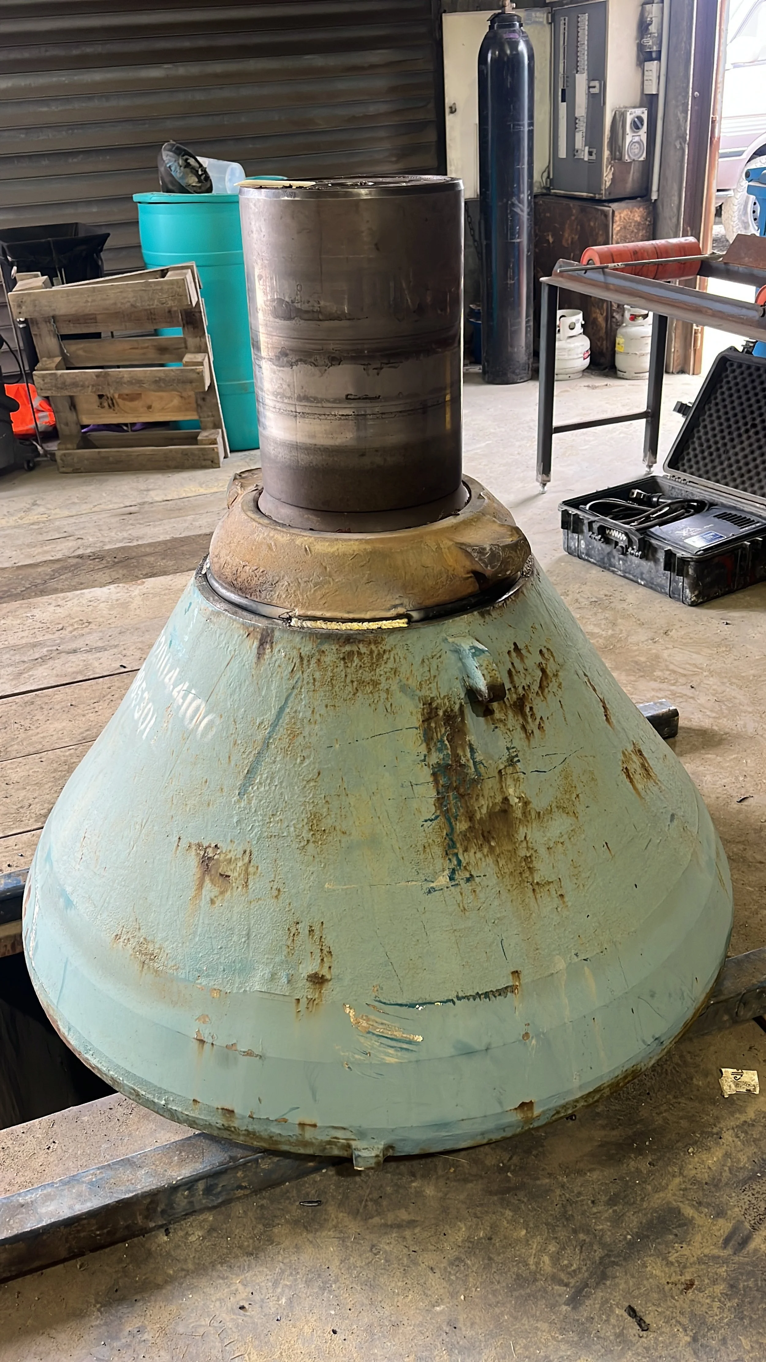 A large, round, metal object with a rusted and worn blue-green surface, sitting in a workshop or garage. On top, there's a black, cylindrical container or pipe. Various tools, equipment, and supplies are visible in the background.