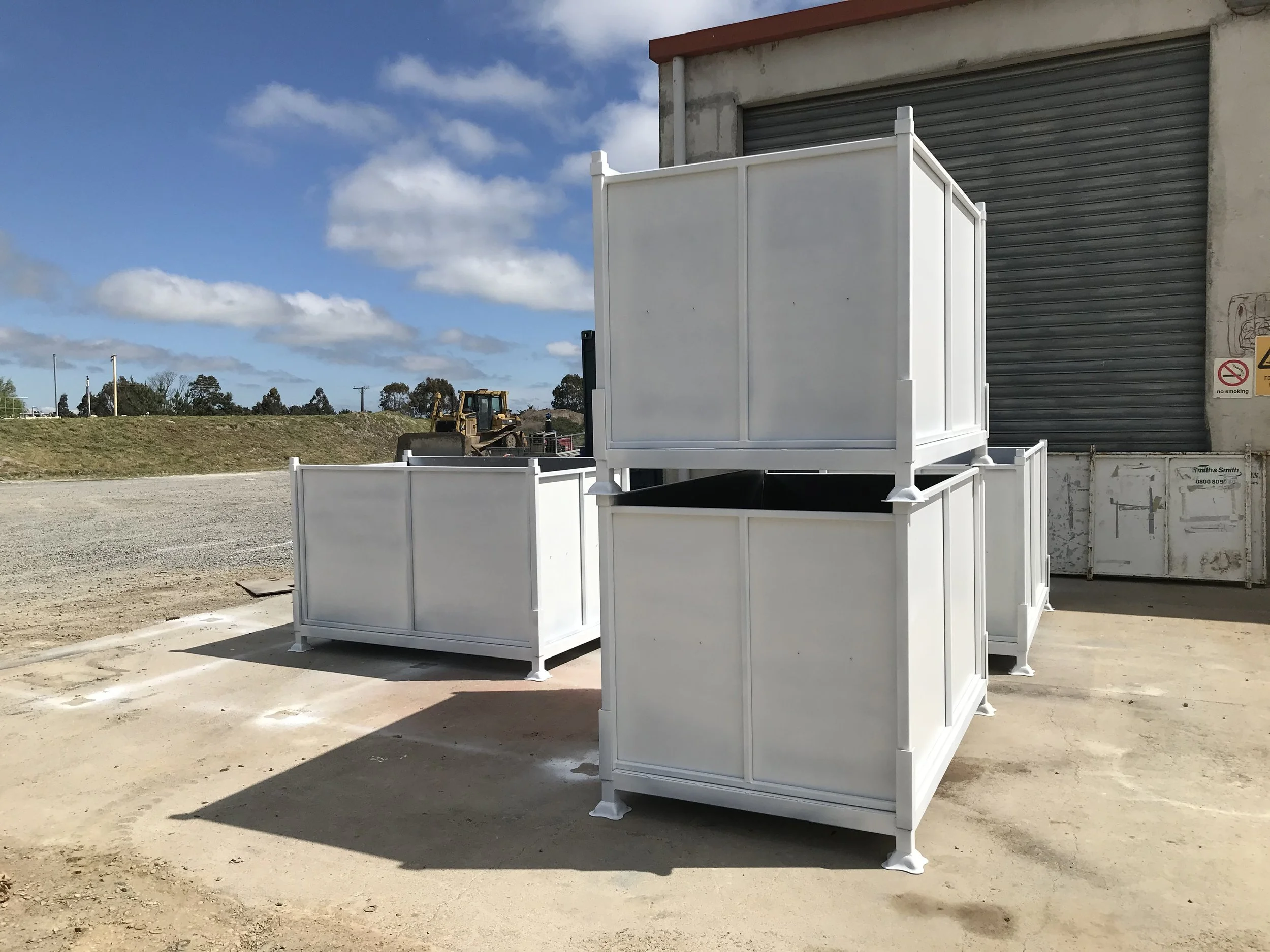 White outdoor storage containers with a partially open top, placed in front of a building with a gray roll-up door, on a gravel surface. Construction equipment and a grassy hill are visible in the background under a partly cloudy sky.