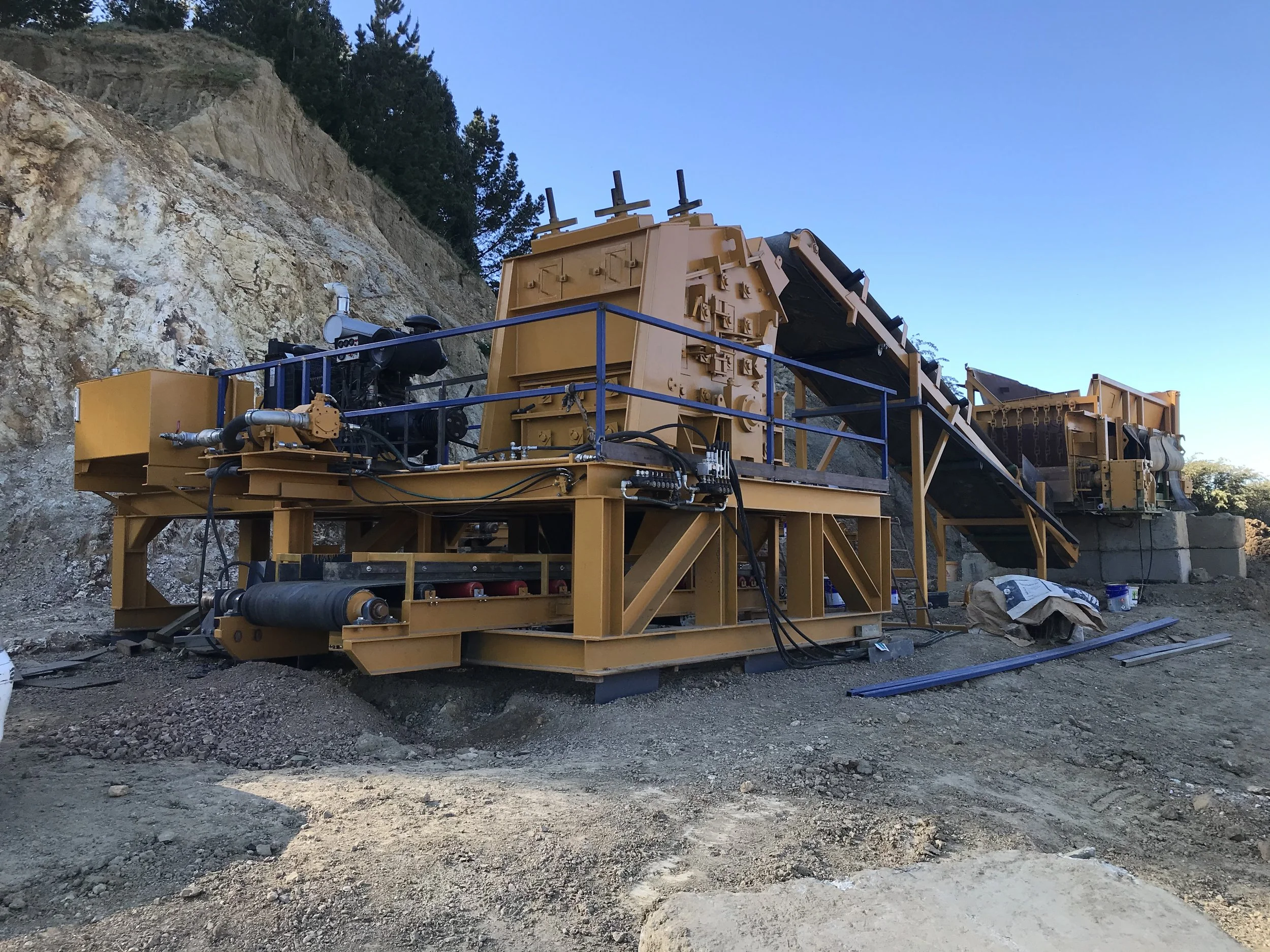 Large yellow industrial machinery at a construction site, with a conveyor belt and mechanical components, set against a dirt landscape and hillside with trees.