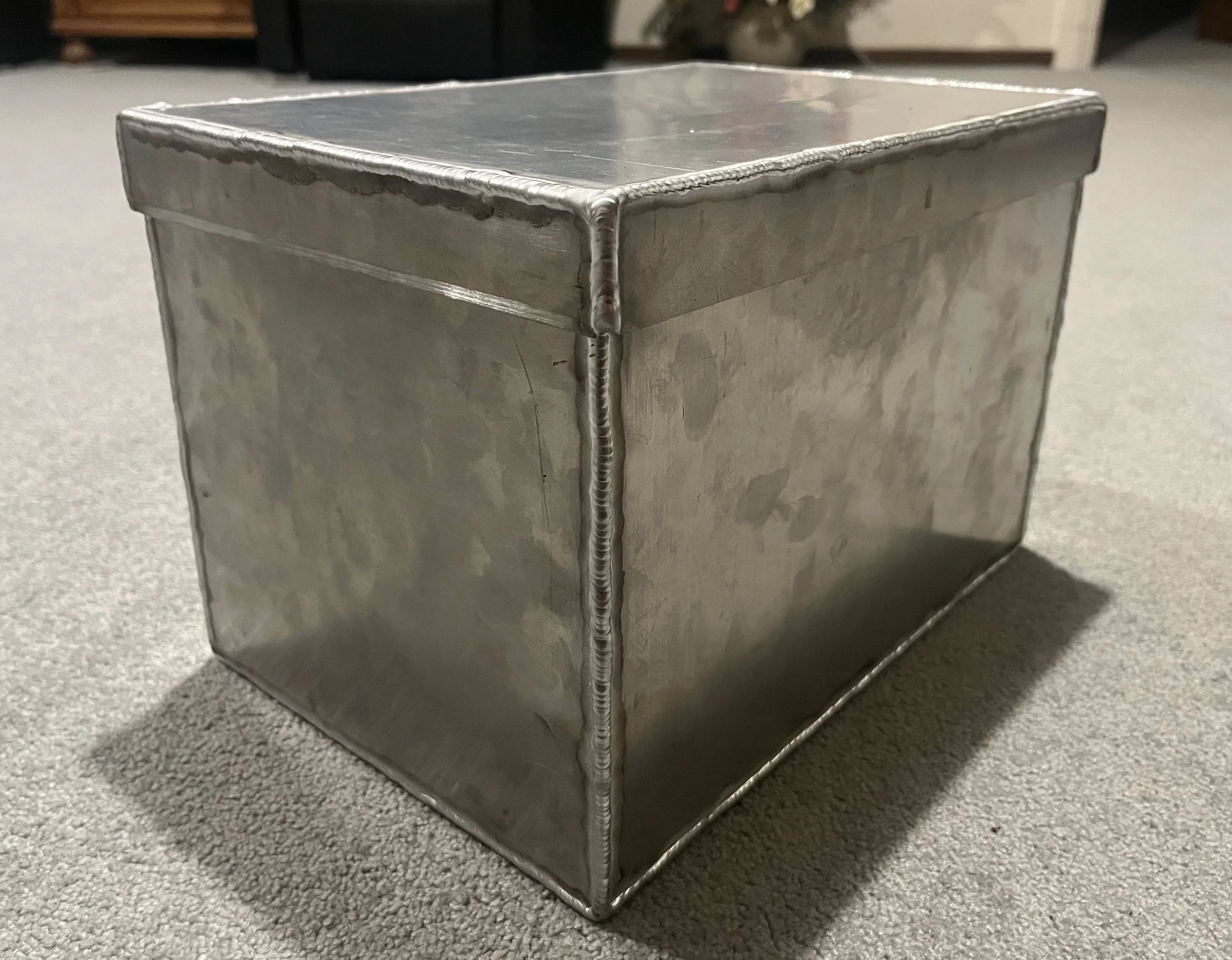 A metal cube with visible welds at the edges, sitting on a carpeted floor with a blurred background.