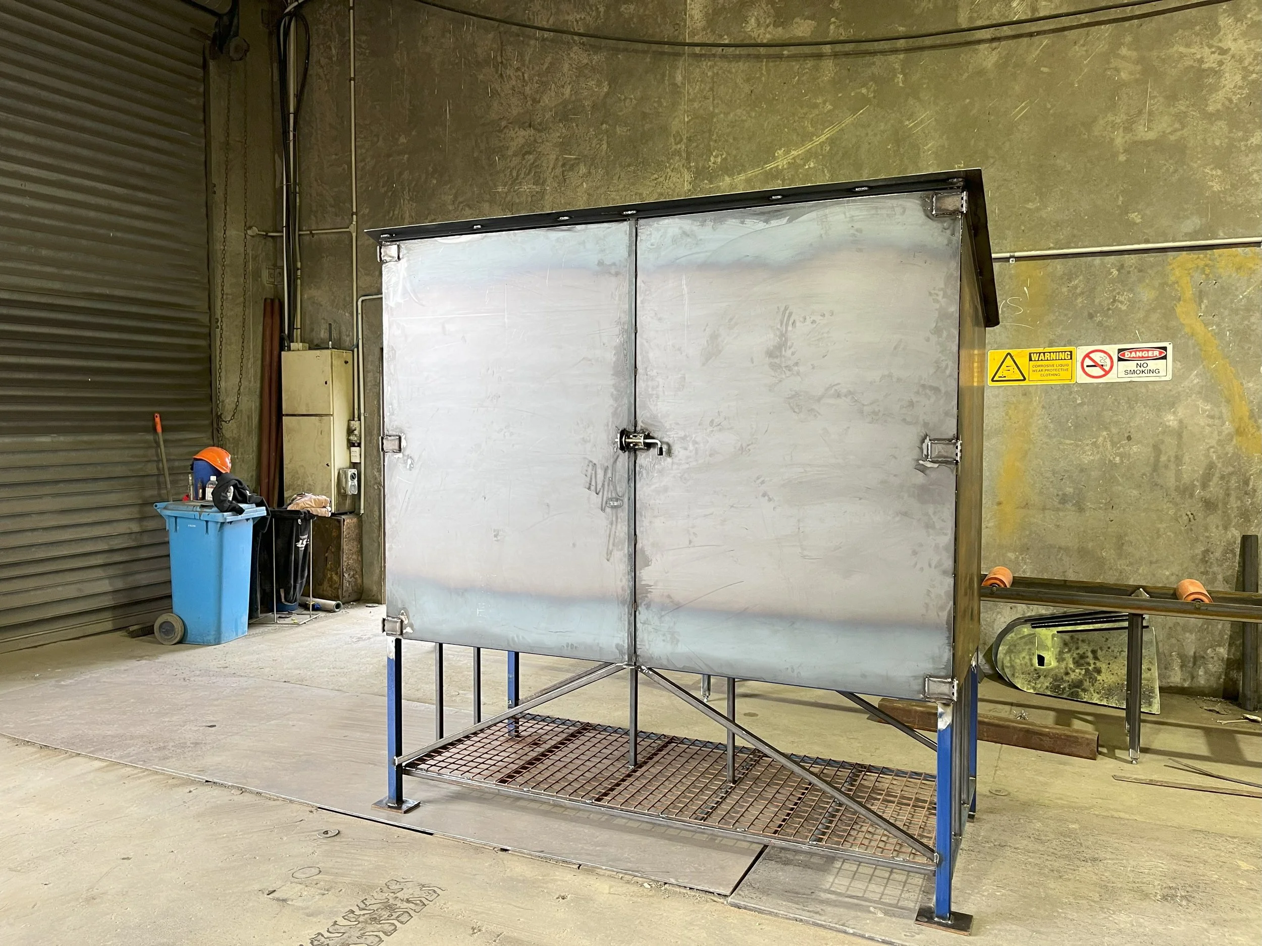 A metal loading rack with a grated platform and a metallic enclosure in an industrial or warehouse setting, with signs warning of dangers and prohibiting smoking on a wall behind it.