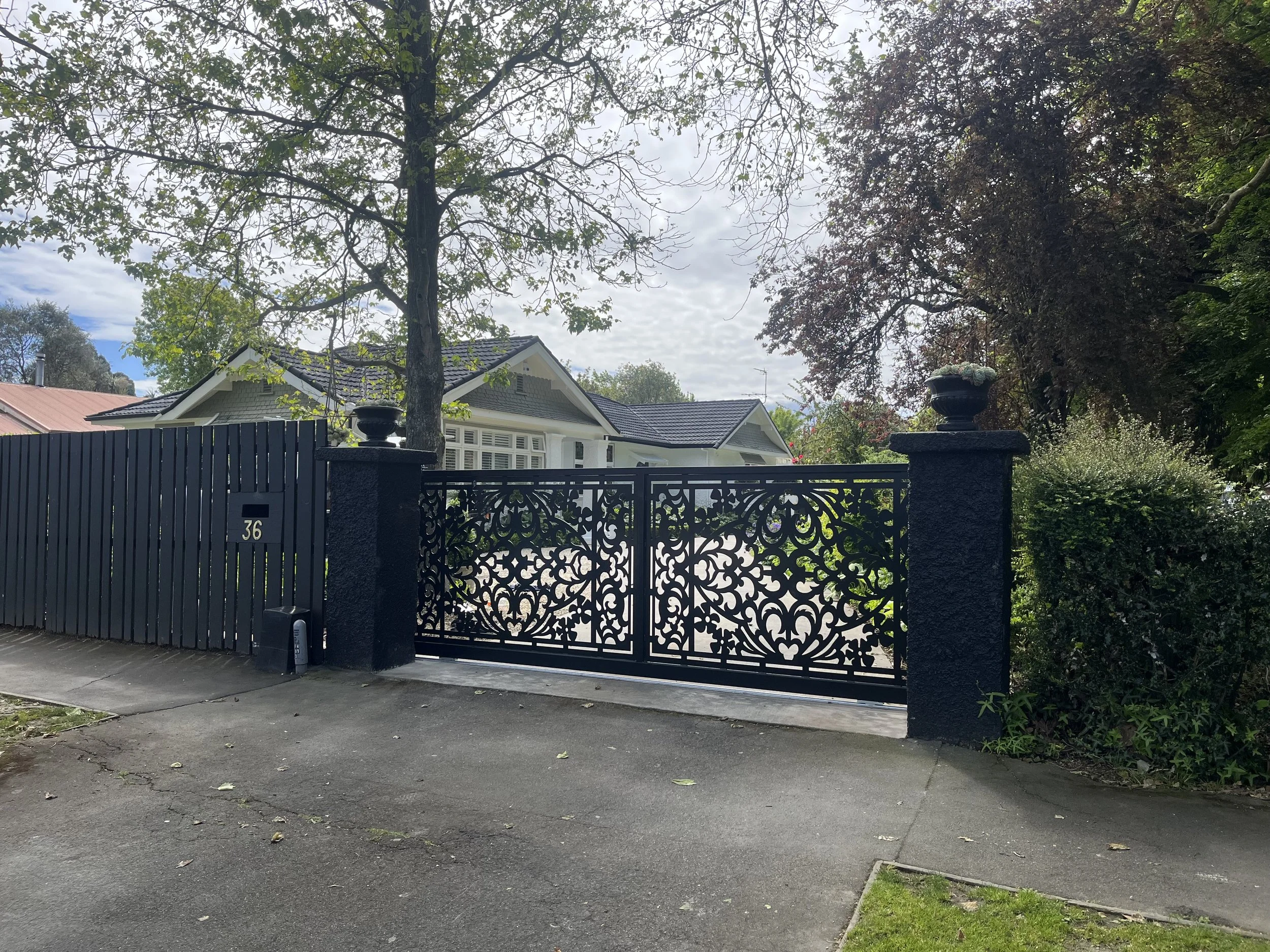 View of a black decorative metal gate with house and trees in the background, cloudy sky overhead, and a small sidewalk and driveway in front.