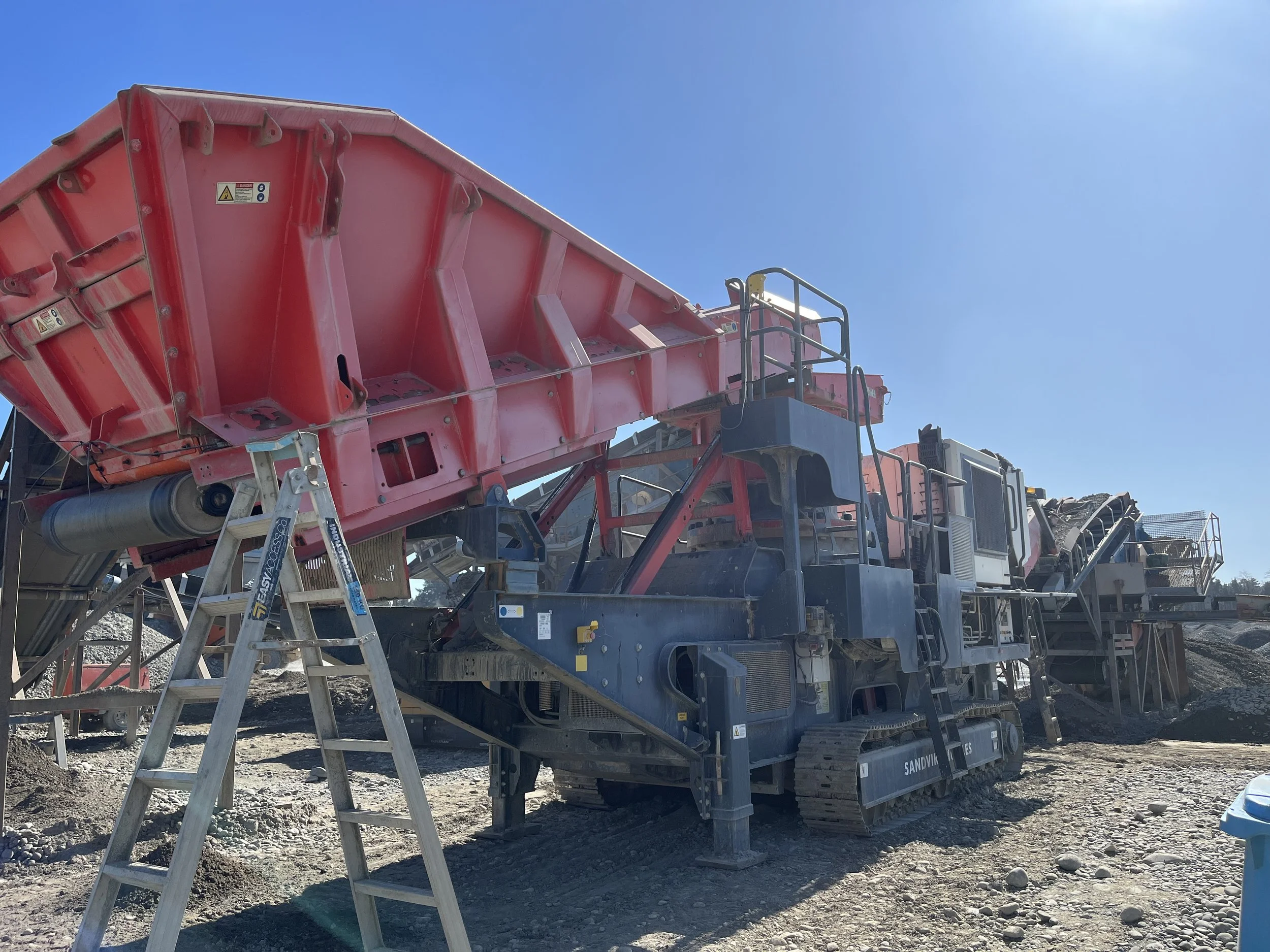 Construction site with a large red mobile crusher machine, a metal ladder, and a blue sky.