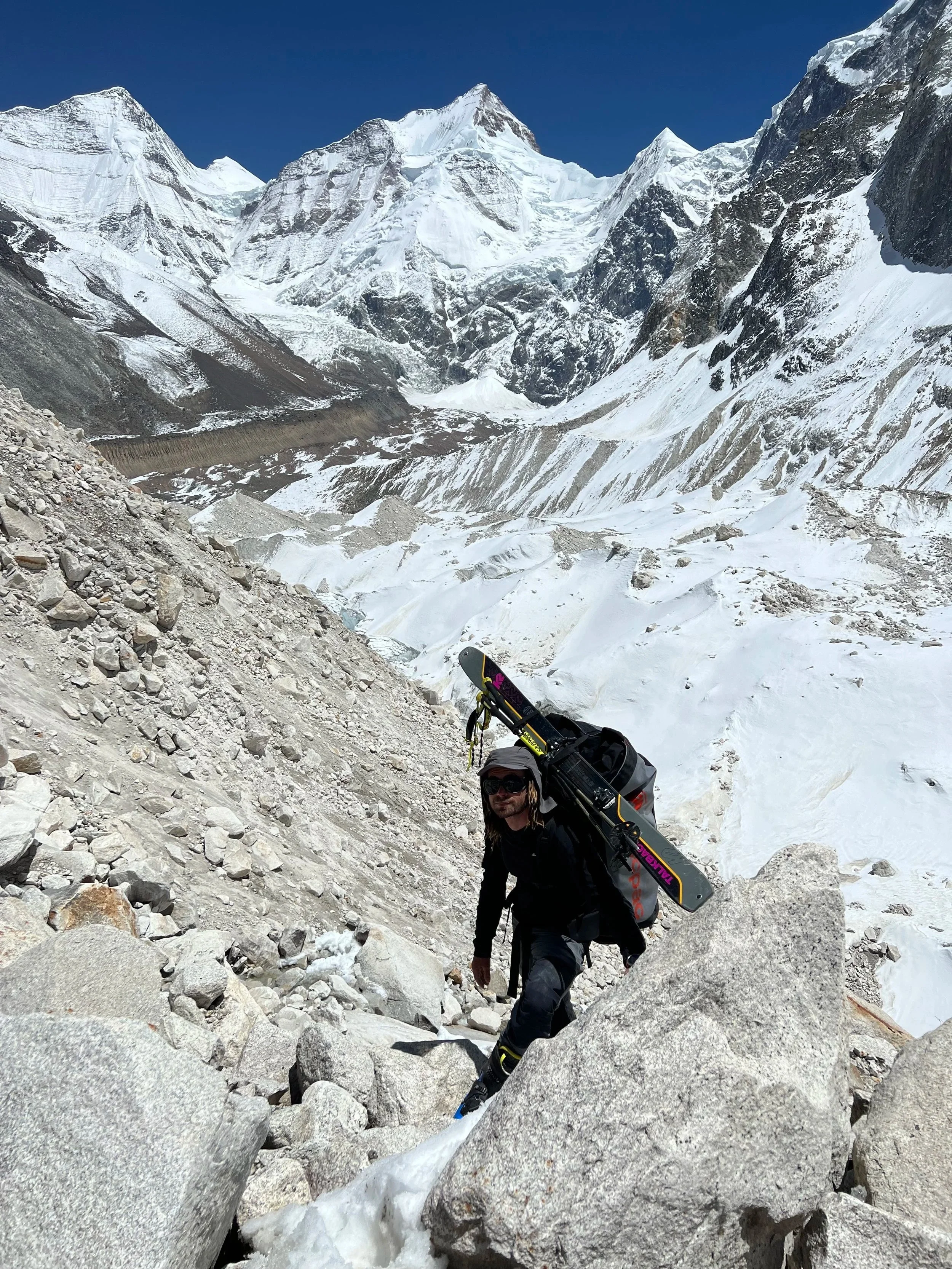 A hiker wearing sunglasses and dark clothing, carrying skis on his shoulder, ascending a rocky, icy slope in a snow-covered mountain landscape with towering peaks in the background.
