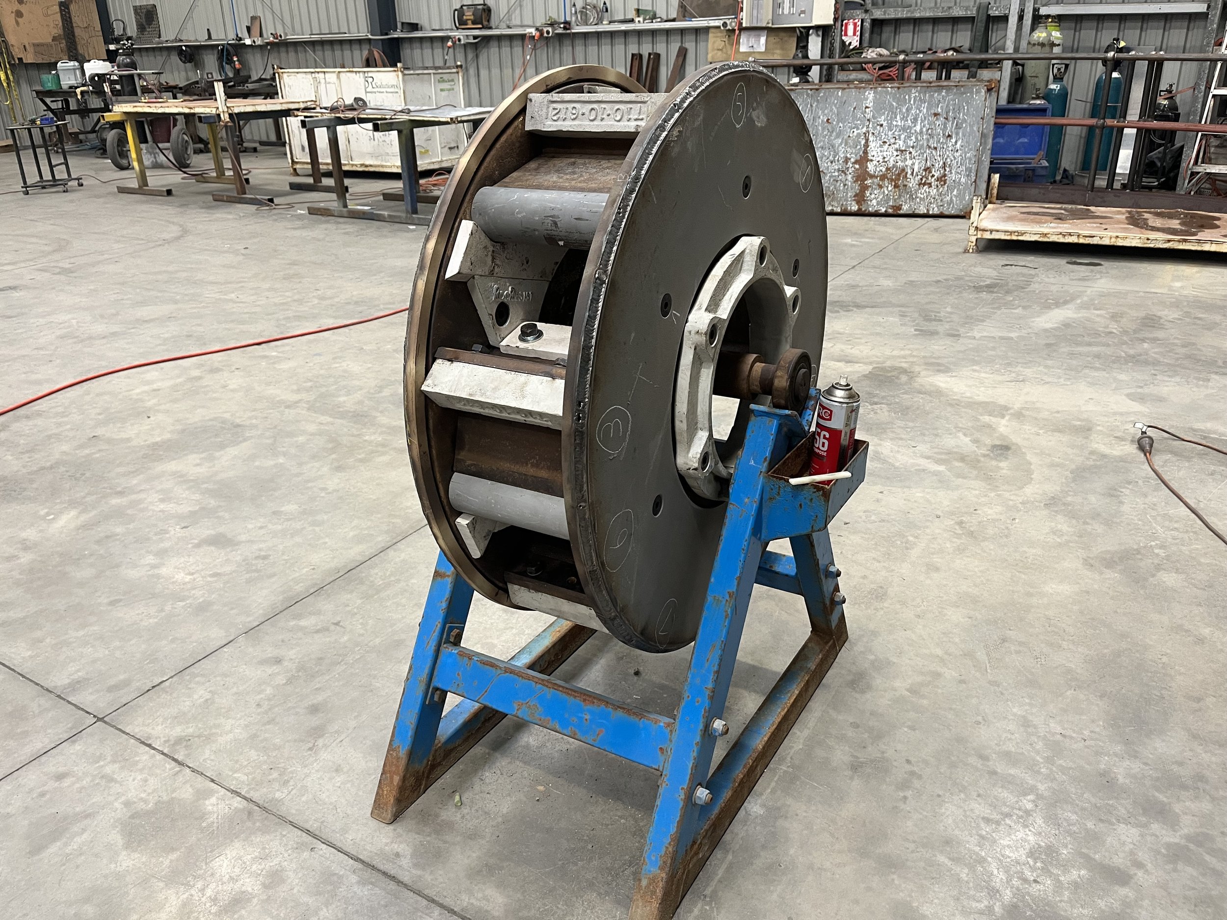Large industrial wheel or pulley mounted on a blue stand in a workshop or warehouse, with various tools and equipment in the background.