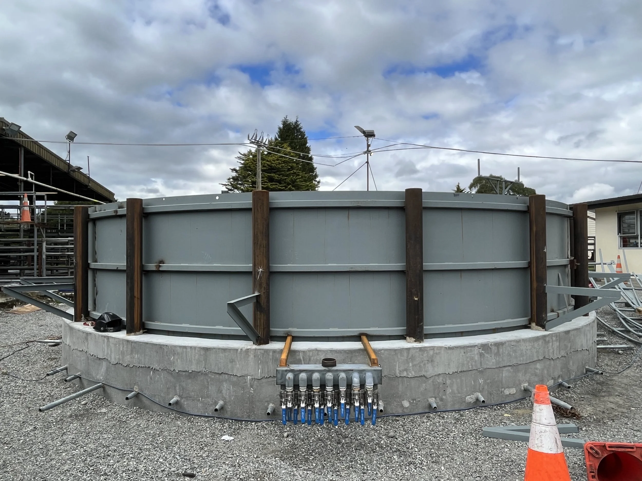 Construction site with a large circular metal tank, scaffolding, electrical wires, and orange safety cones and barrier.