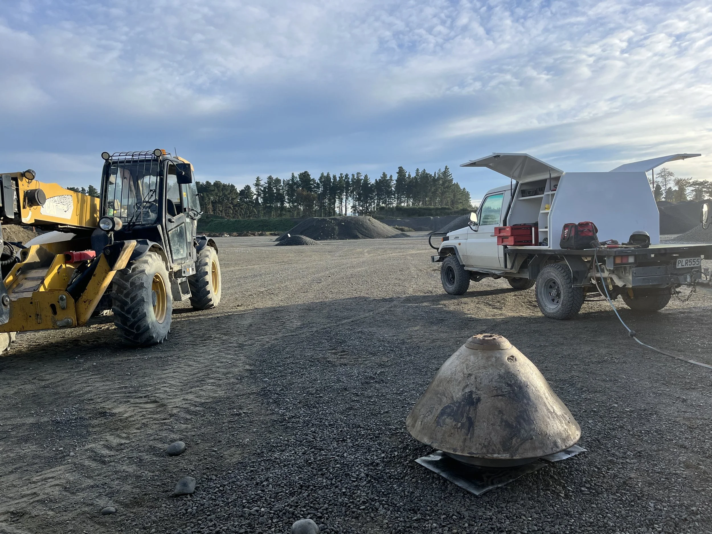 Construction site with a cement mixer, a yellow heavy machinery vehicle, and a white utility truck with the hatch open, surrounded by gravel and dirt, with trees and a partly cloudy sky in the background.
