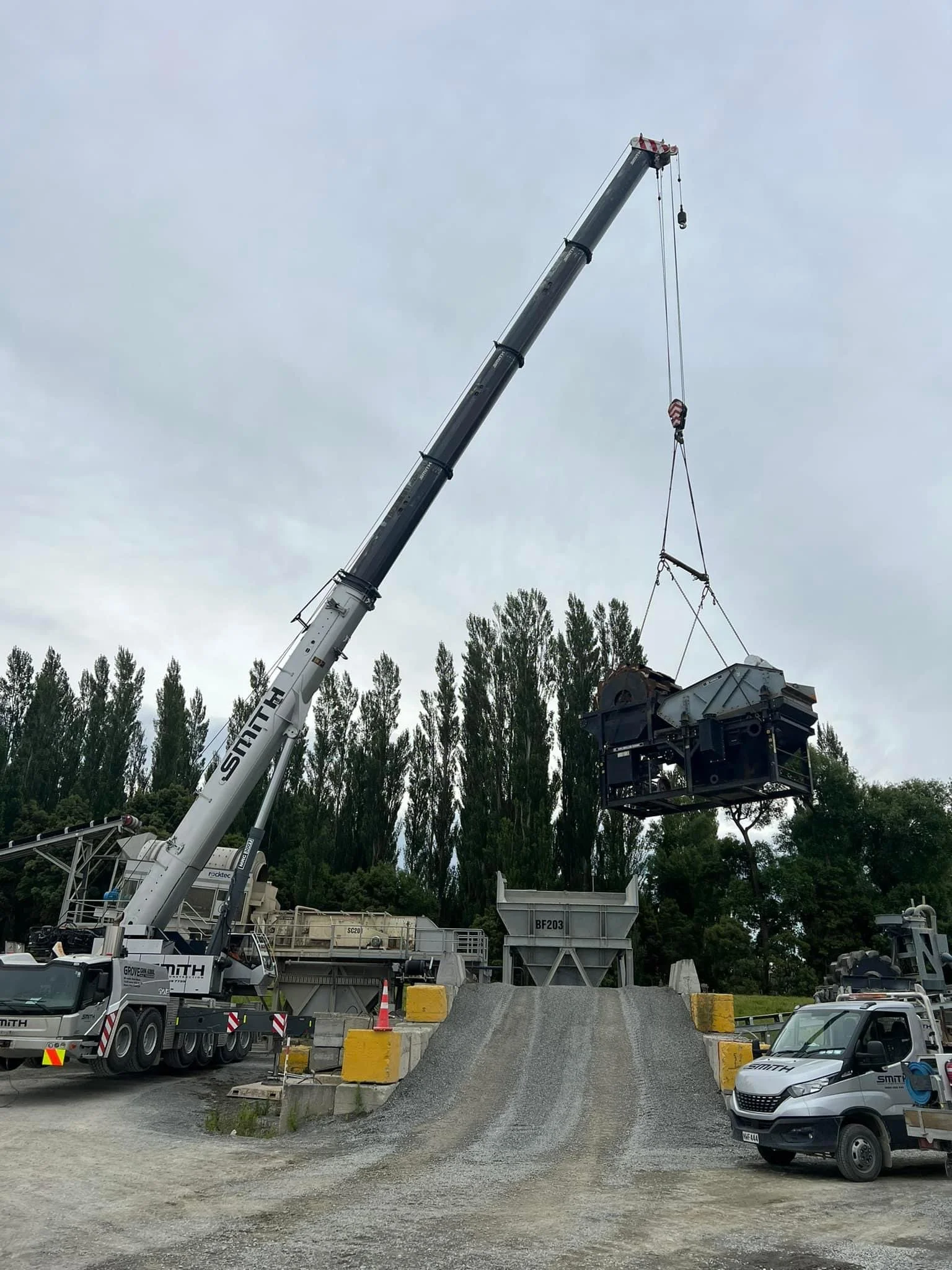 A construction crane lifting a large load onto a truck at a construction site with trees in the background and overcast sky.