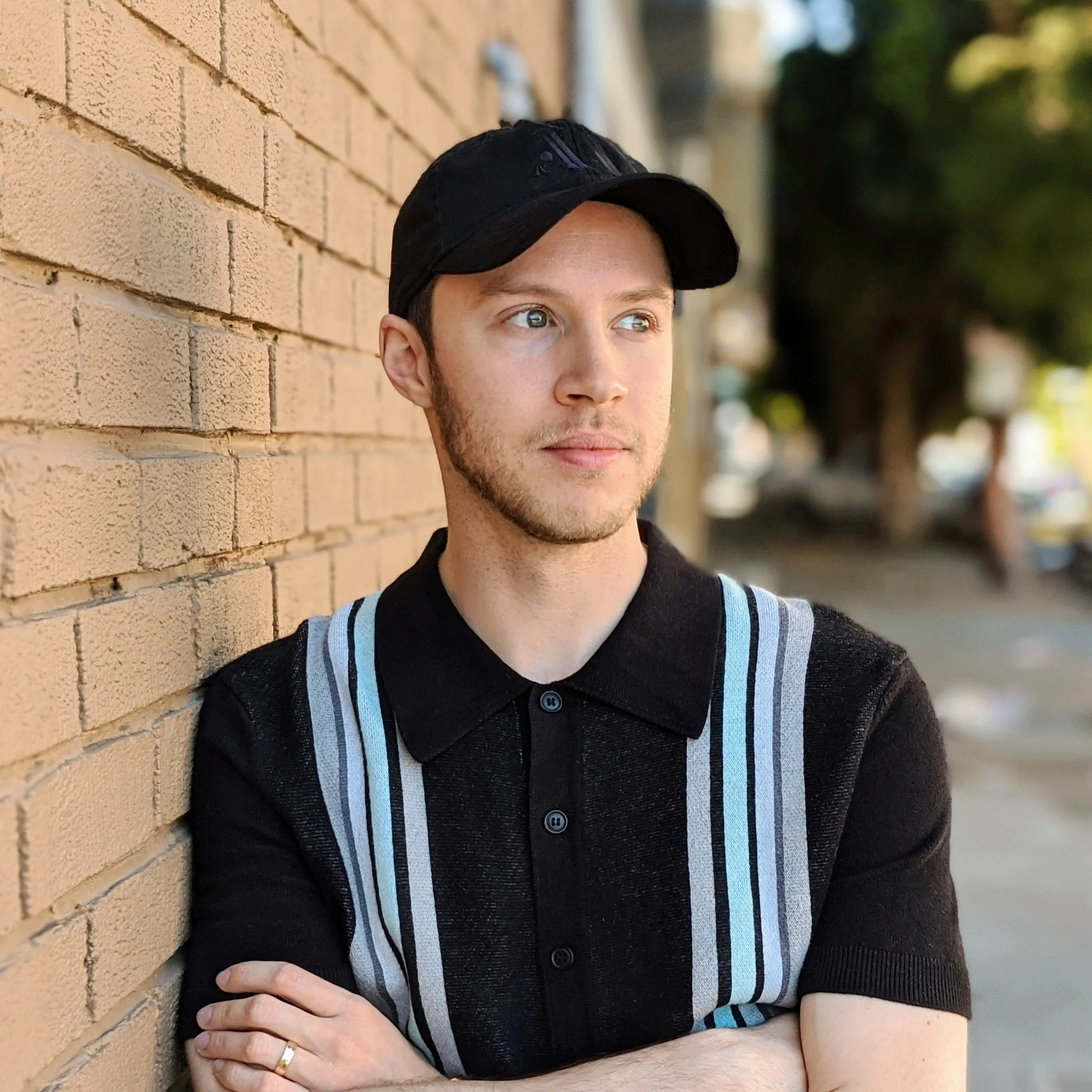A young man with light skin, blue eyes, and short dark hair wearing a black cap, a black shirt, and a black sweater with blue and white stripes on the sleeves, standing outdoors against a brick wall with blurred trees in the background.