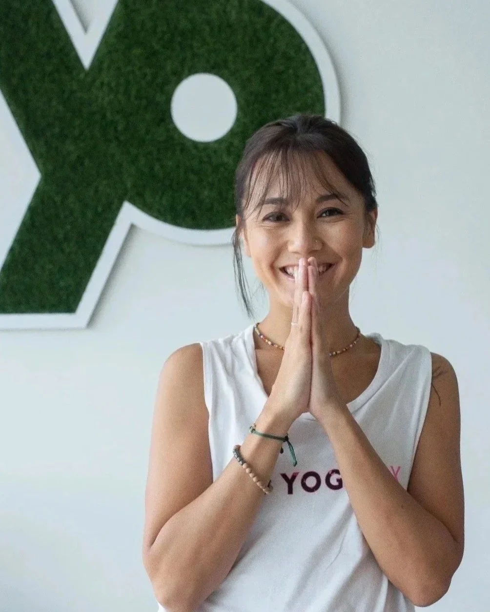 A smiling woman with dark hair, wearing a white sleeveless shirt with the word "YOGA" on it, standing in front of a white wall with a large green letter P behind her, holding her hands together in a prayer gesture.