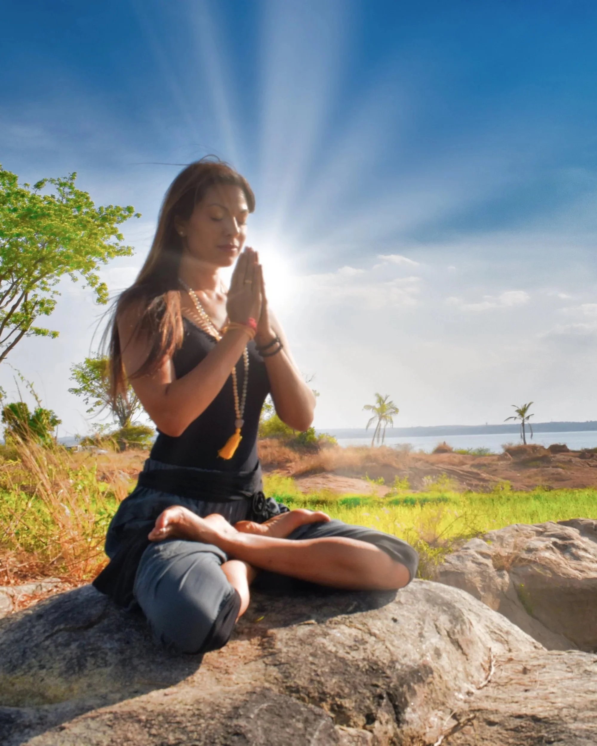 A woman practicing yoga outdoors on a rock, sitting in a meditative pose with hands in prayer position, wearing black attire and jewelry, with a scenic background of trees, water, and a bright sky.