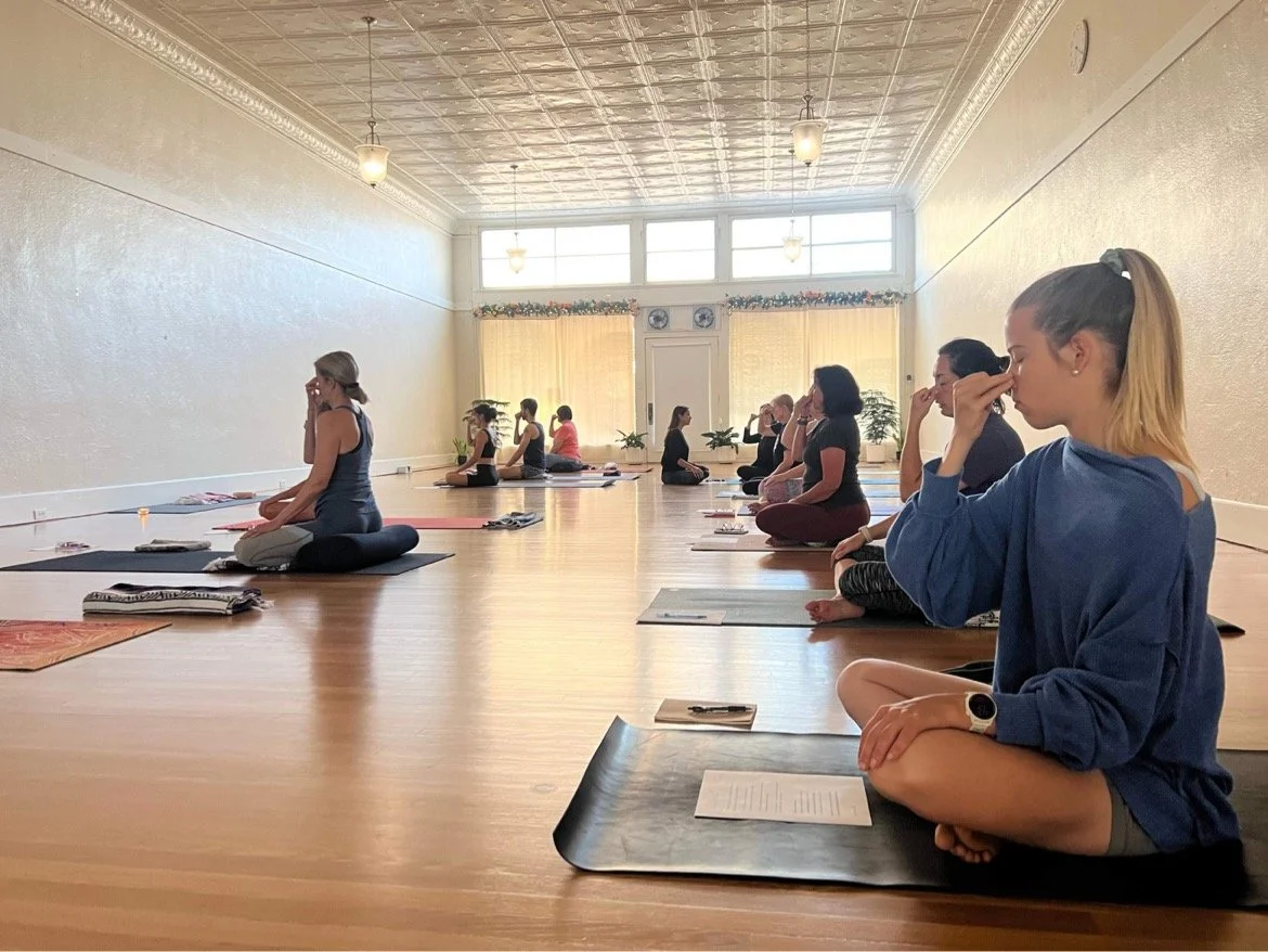 Group of people practicing yoga in a spacious, well-lit studio with large windows, sitting cross-legged on yoga mats, each with personal belongings nearby.