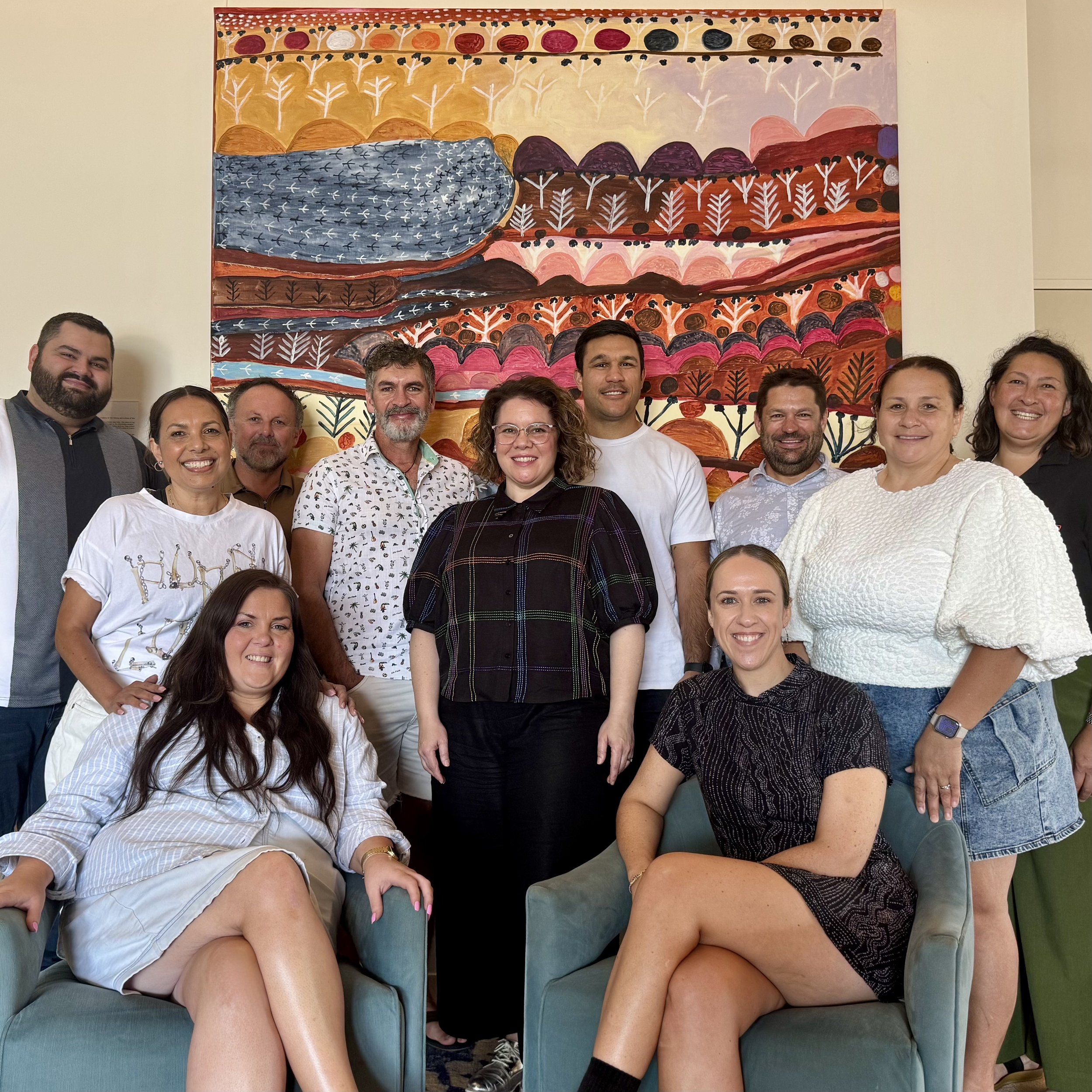 A group of eleven people in a room with a large colorful abstract painting on the wall behind them. The group is smiling and posing for the photo, with some sitting on armchairs and others standing behind them.