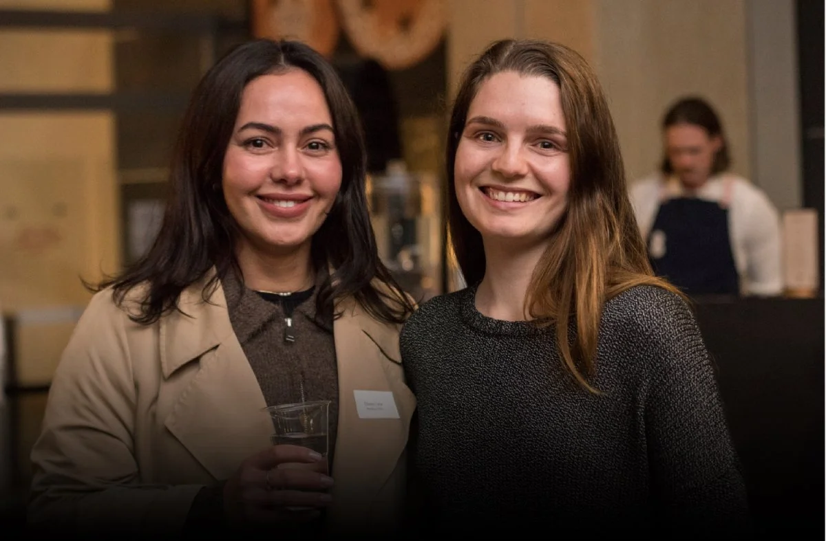 Two women smiling at a social event, standing indoors, one holding a glass of water, with a blurred background and a woman working at a counter in the background.