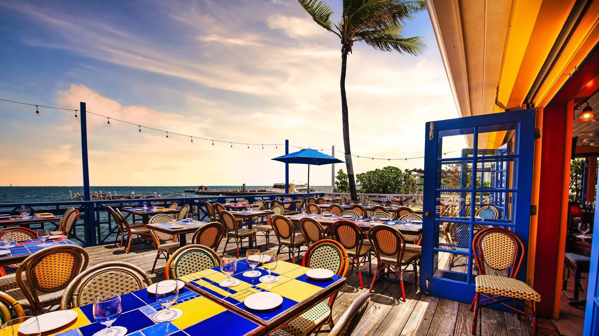 Outdoor restaurant patio with colorful tables and chairs, overlooking the ocean at sunset, with string lights, a blue door, an umbrella, and a palm tree.