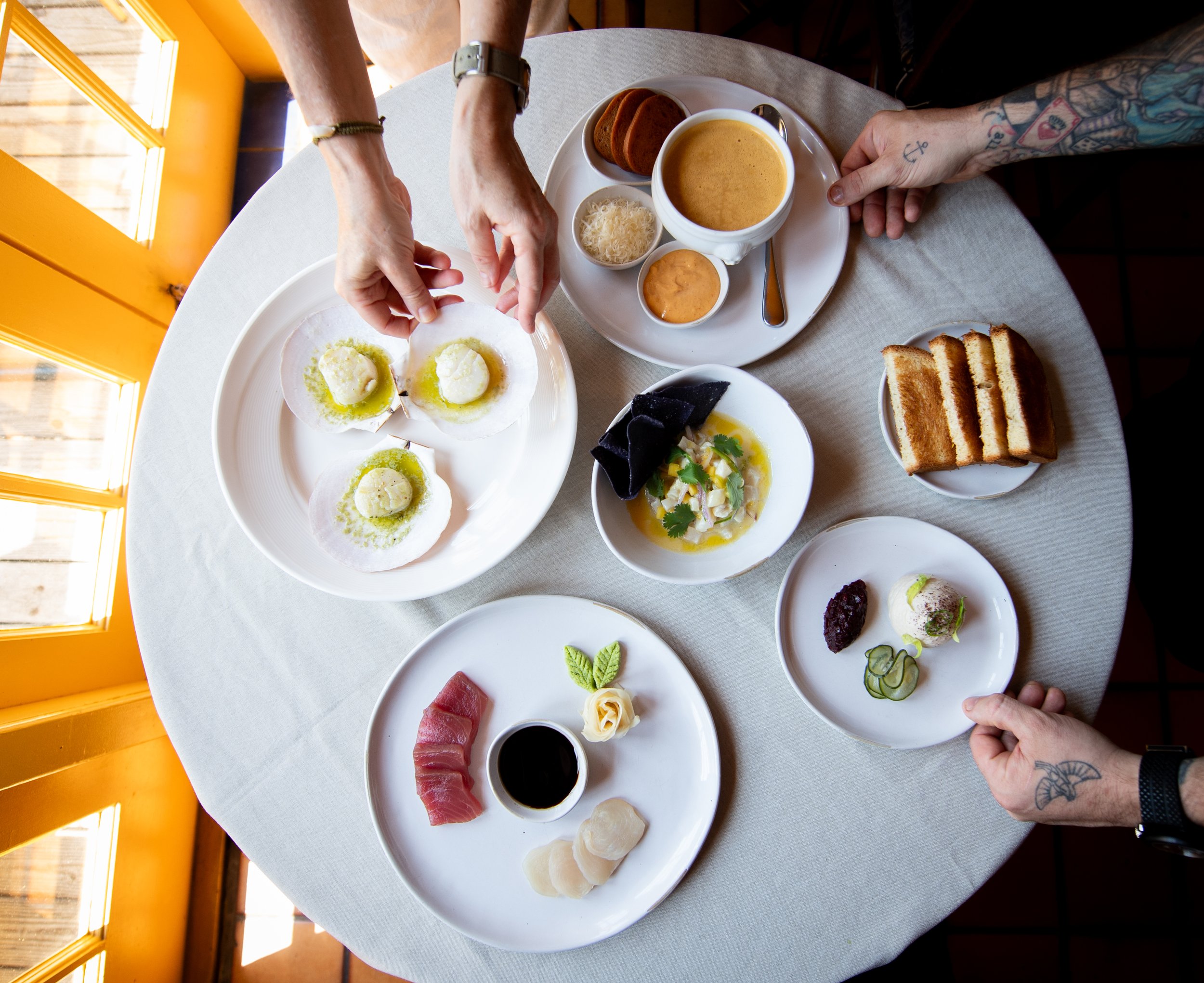 A top-down view of a table set with various dishes, including scallops, a bowl of soup, toast, sashimi, greens, and desserts, with four hands reaching for the food.