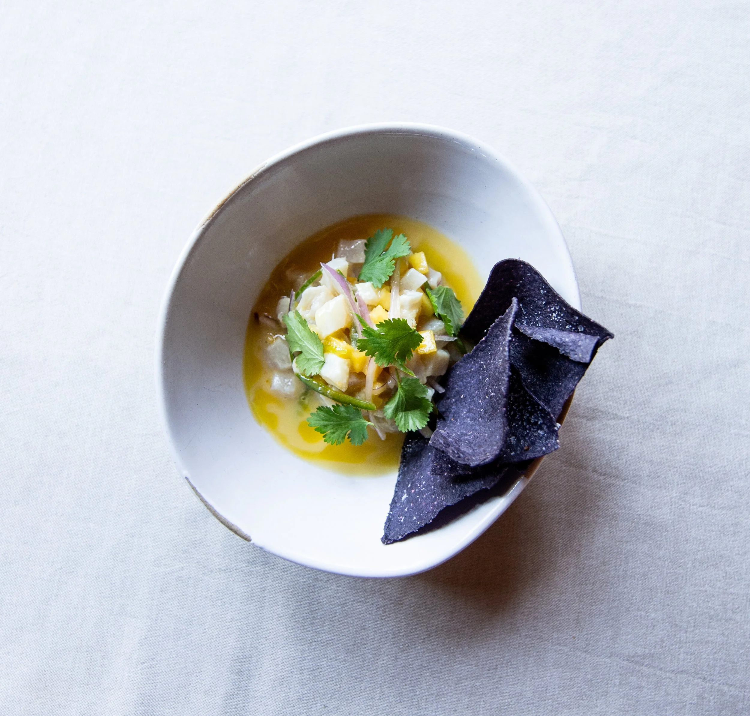 A bowl of ceviche garnished with cilantro and served with blue corn chips across a light-colored table.