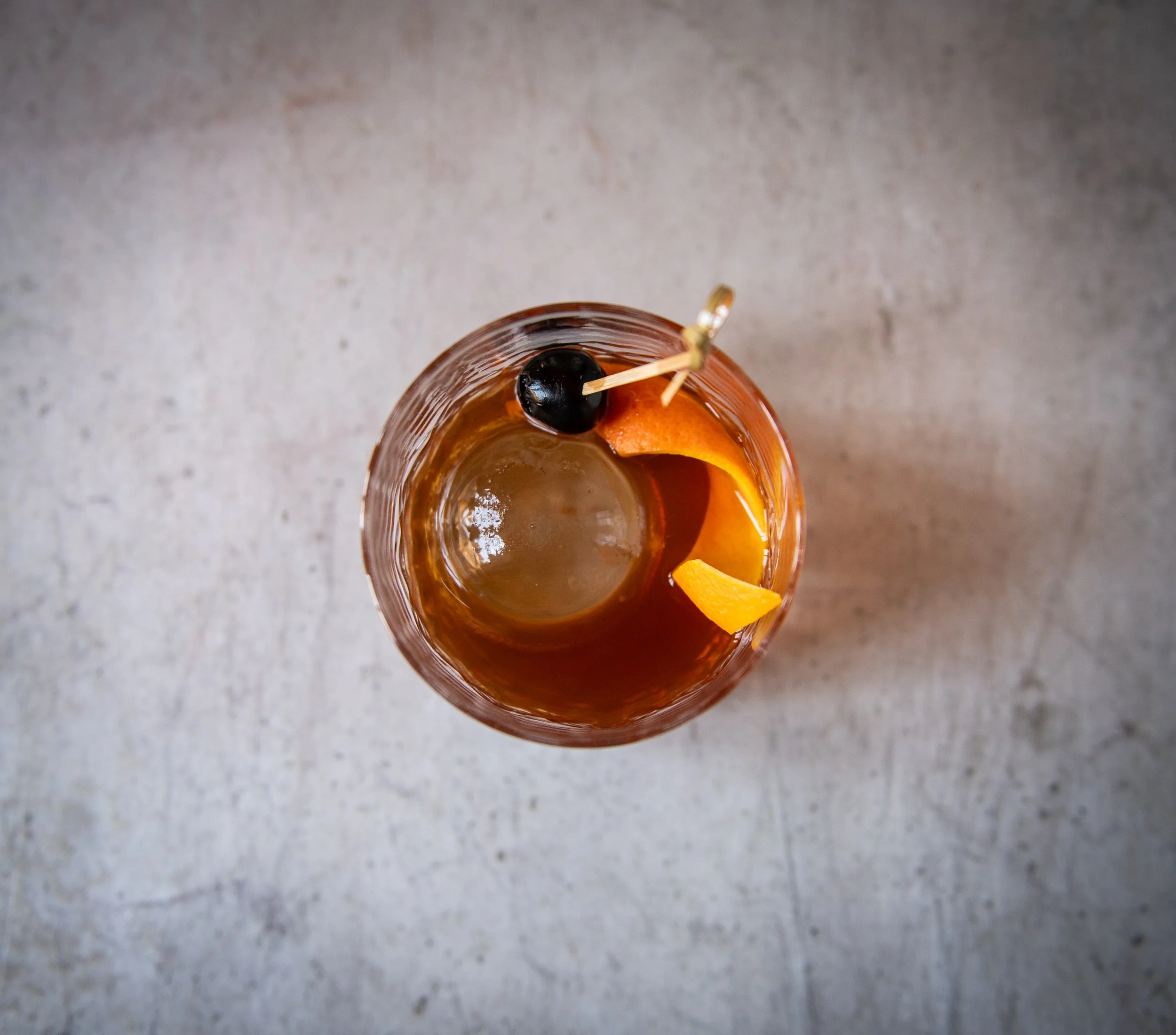 Top-down view of a glass with an amber-colored cocktail, garnished with a black cherry, an orange peel, and a small orange slice. The drink contains ice and is placed on a textured, light gray surface.