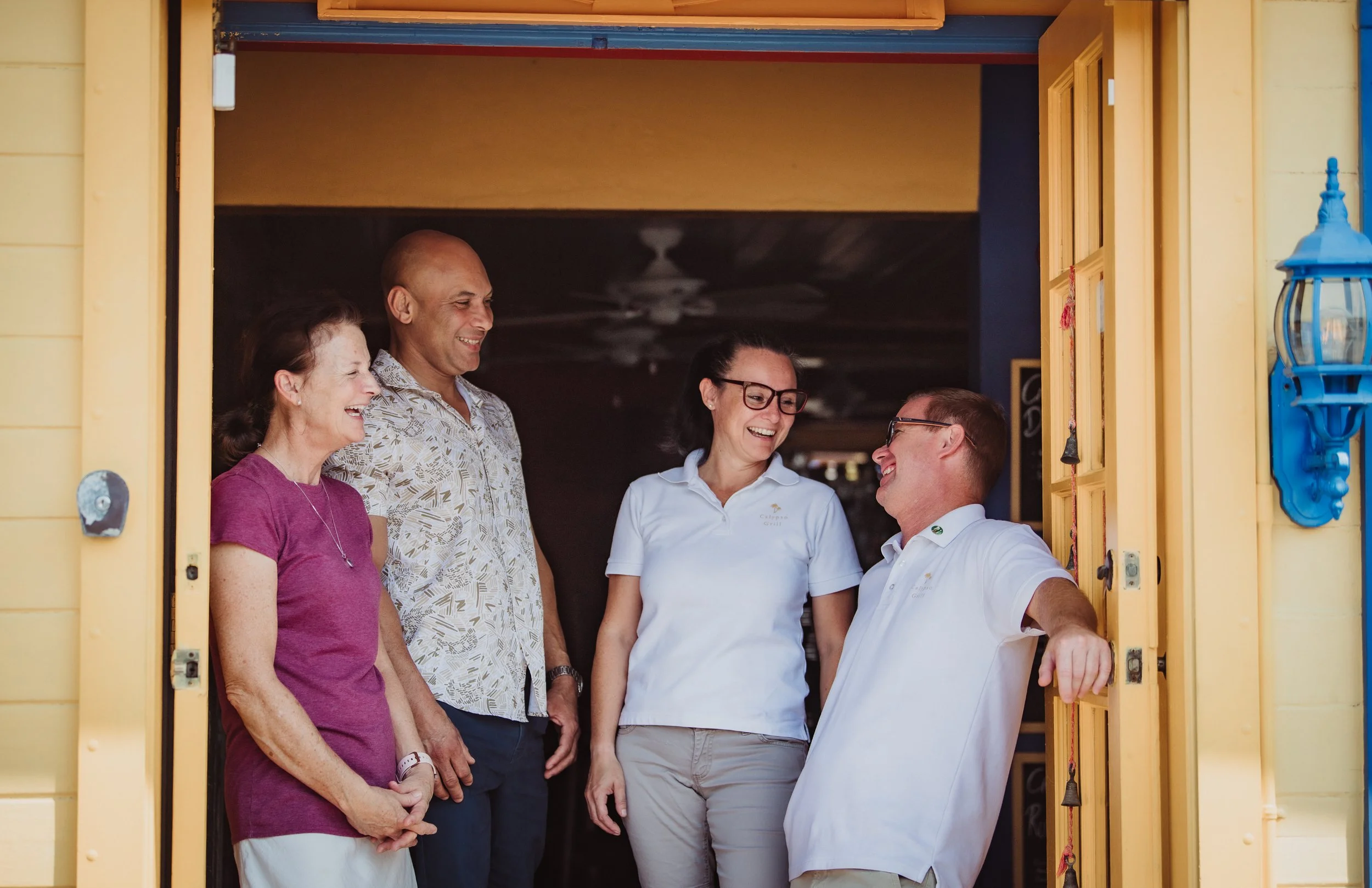 Five people standing at a doorway, engaged in conversation and smiling, with a yellow exterior wall and a blue light fixture visible.