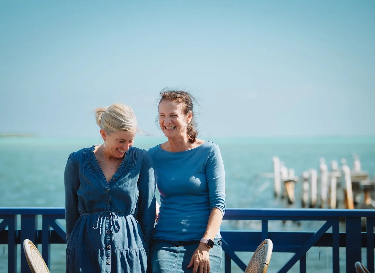 Two women smiling and talking on a balcony overlooking the ocean on a sunny day.